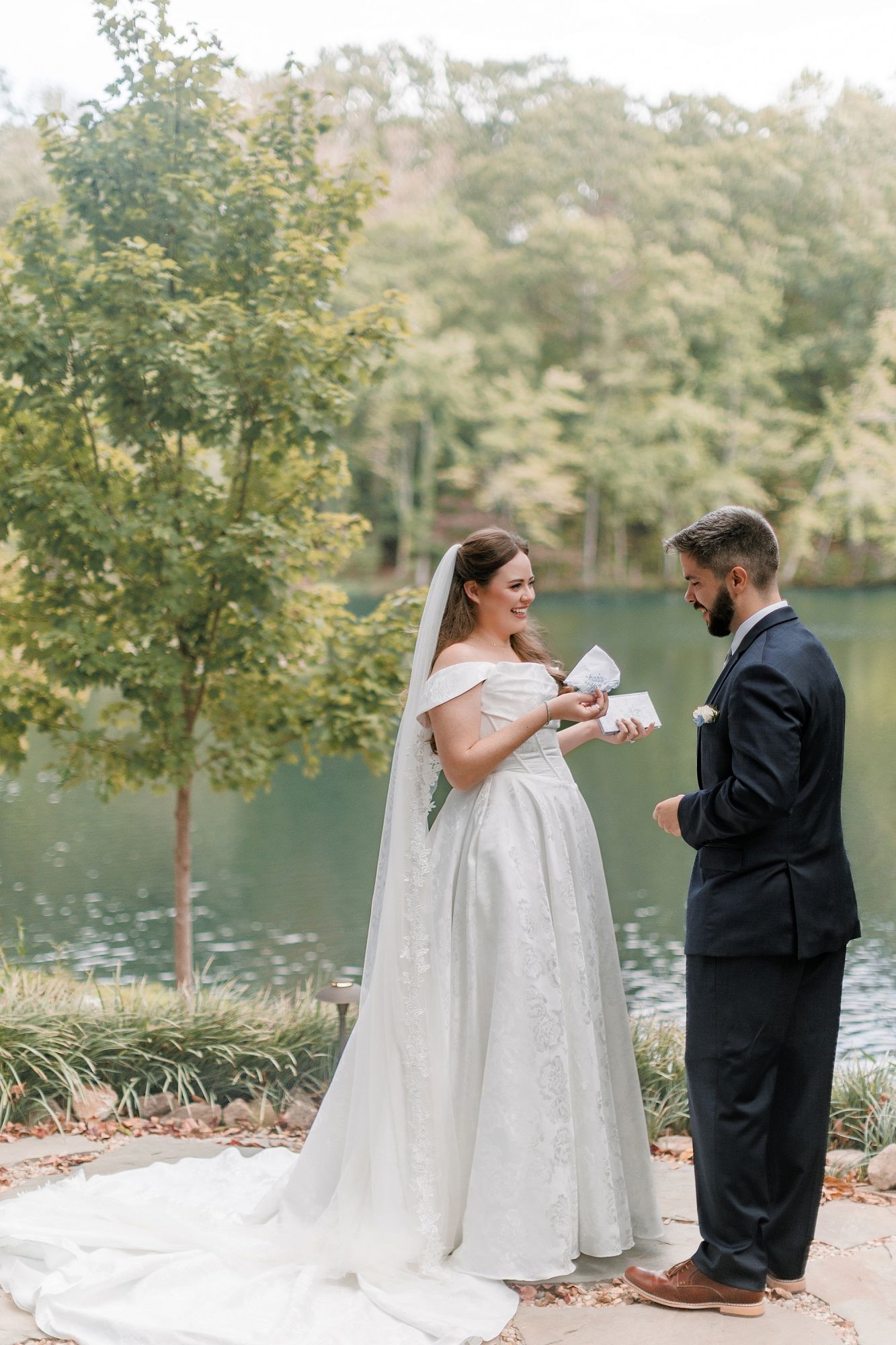Bride in white gown and groom in navy suit exchange vows by a serene lake surrounded by lush green trees.