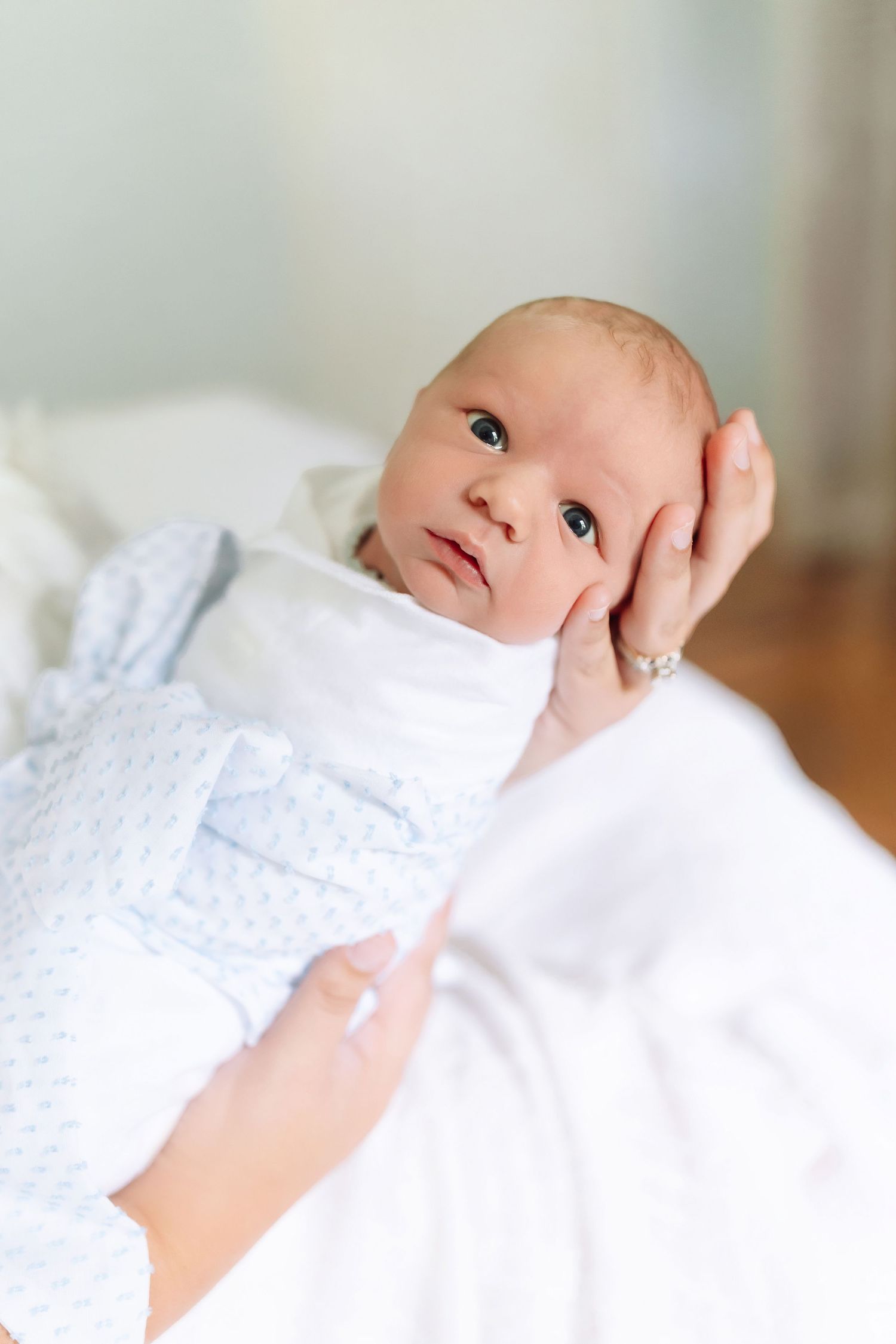 Newborn baby wrapped in white blanket being gently cradled in mother's hands, looking up with alert dark eyes.