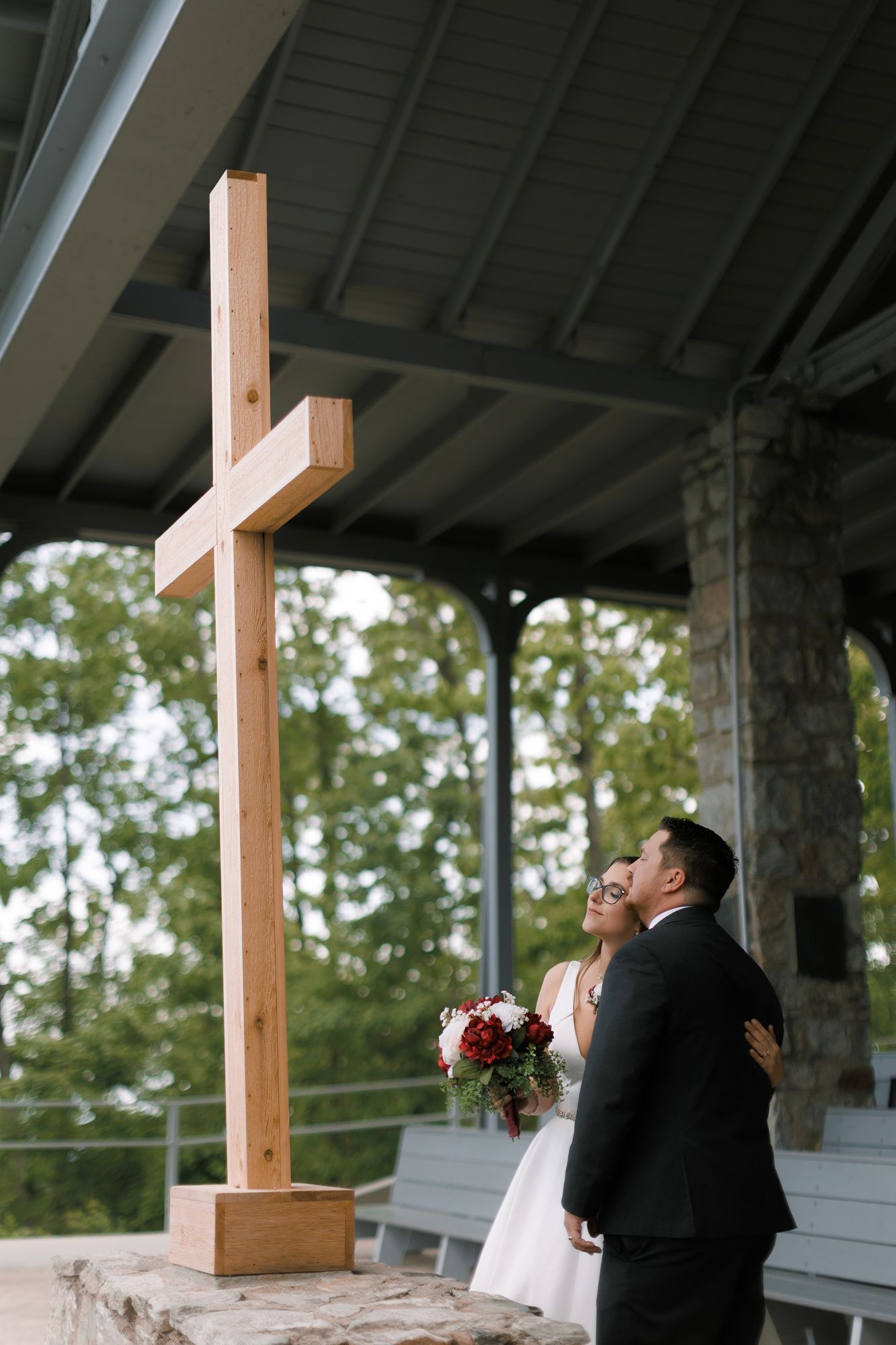 Bride and groom embracing under a wooden cross at an outdoor chapel wedding ceremony venue.