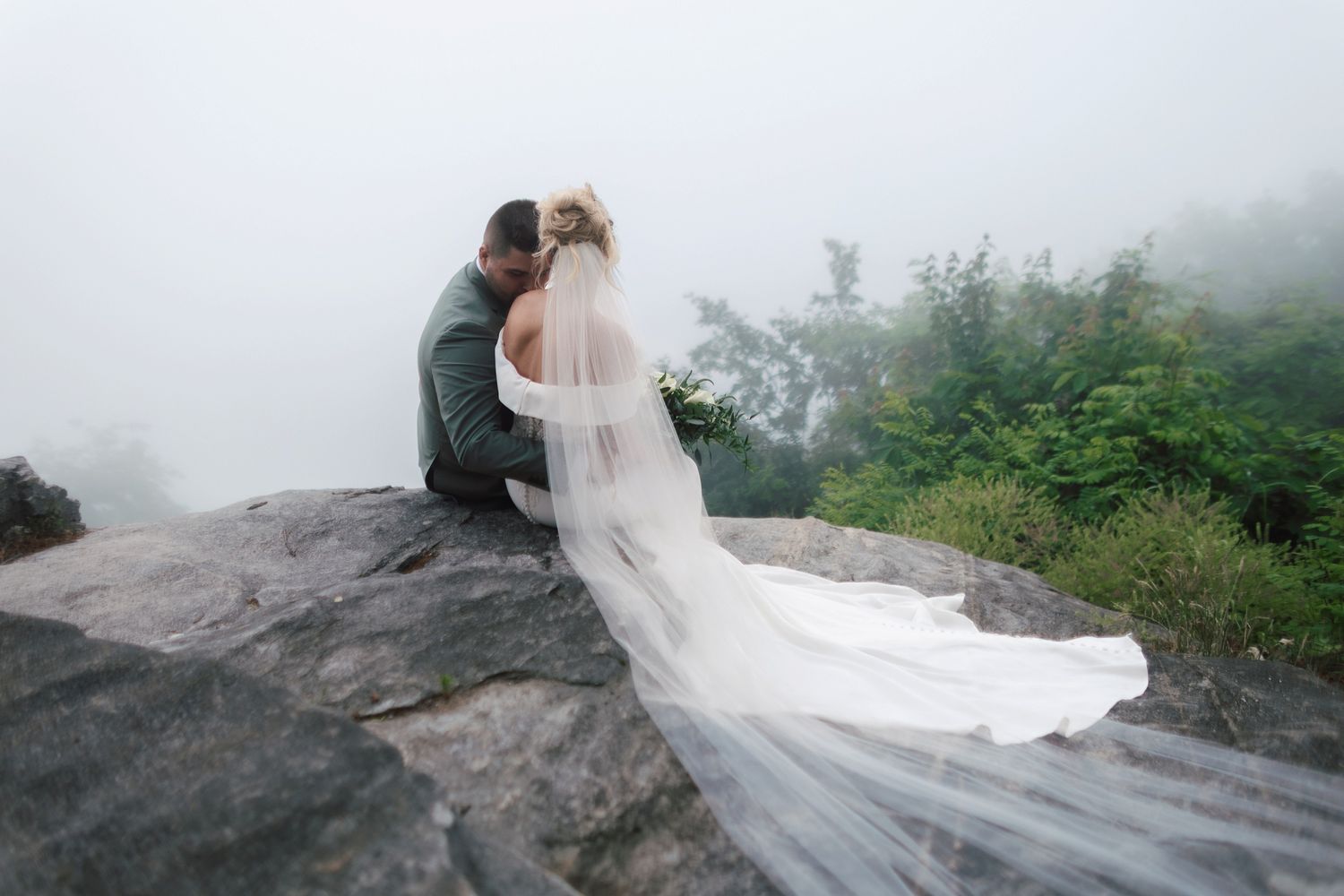 Bride and groom sit intimately on a rocky mountain summit surrounded by misty fog with veil trailing behind.