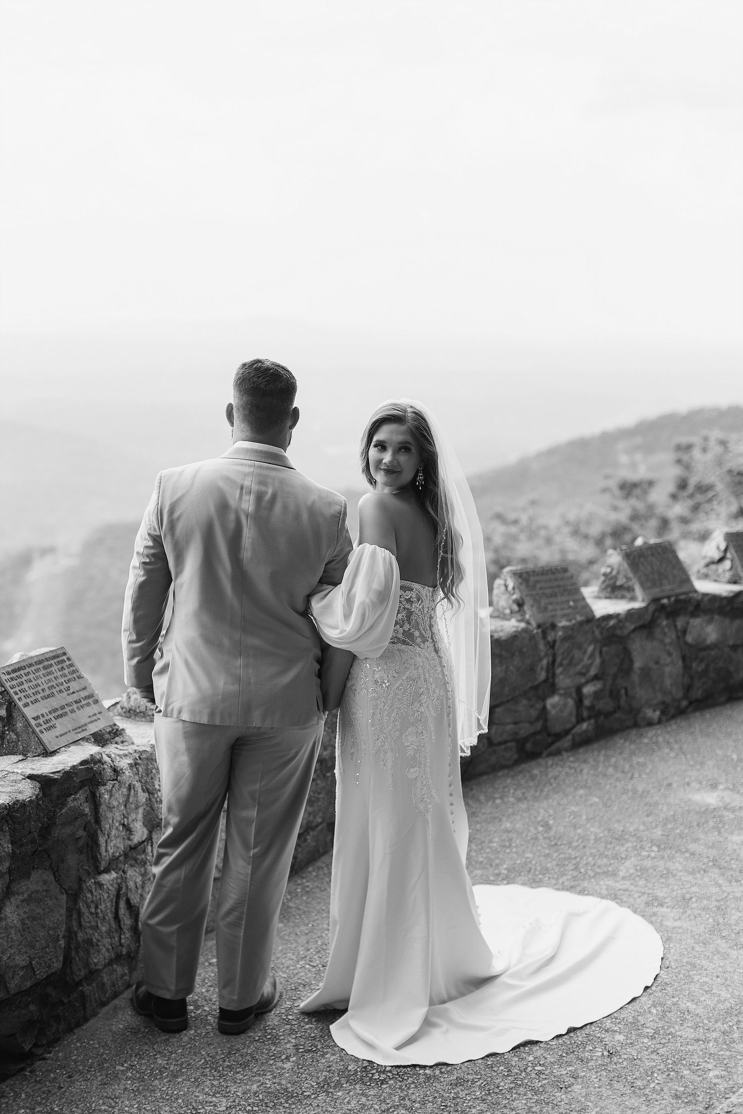 Black and white wedding photo showing a couple standing at a stone wall overlooking mountains.