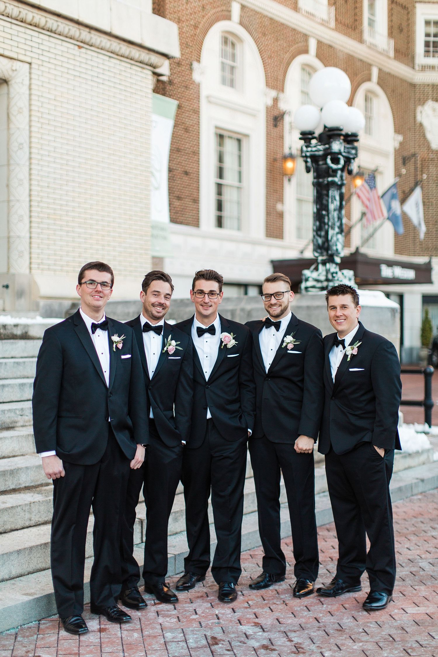 Groomsmen in black tuxedos pose on brick steps outside a historic building.