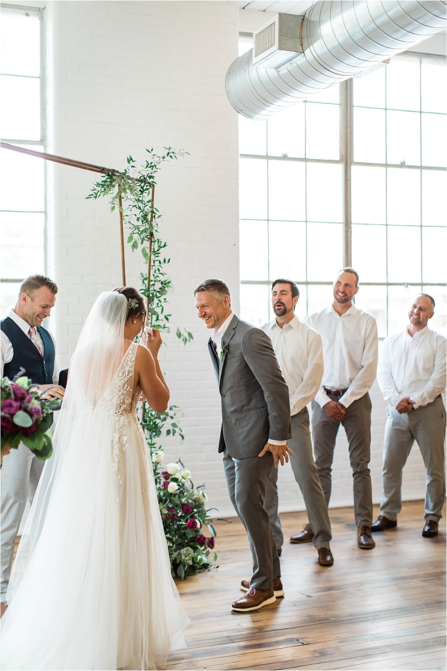 Wedding party stands at altar with copper arch and greenery in bright industrial venue.