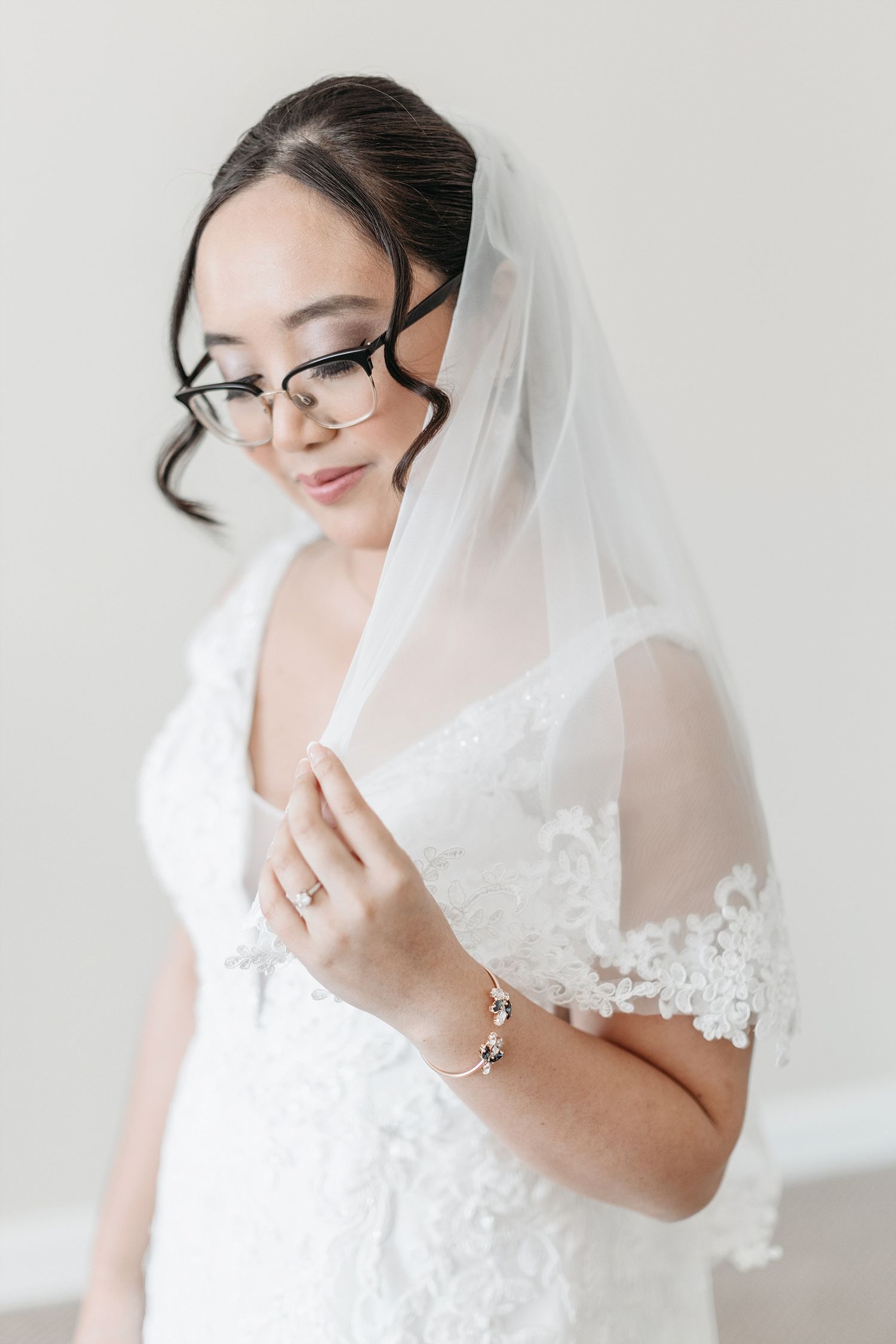A bride in glasses and veil poses elegantly in a white lace wedding gown.