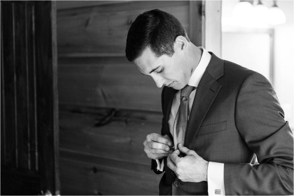 A groom in a dark suit adjusts his tie while getting ready.