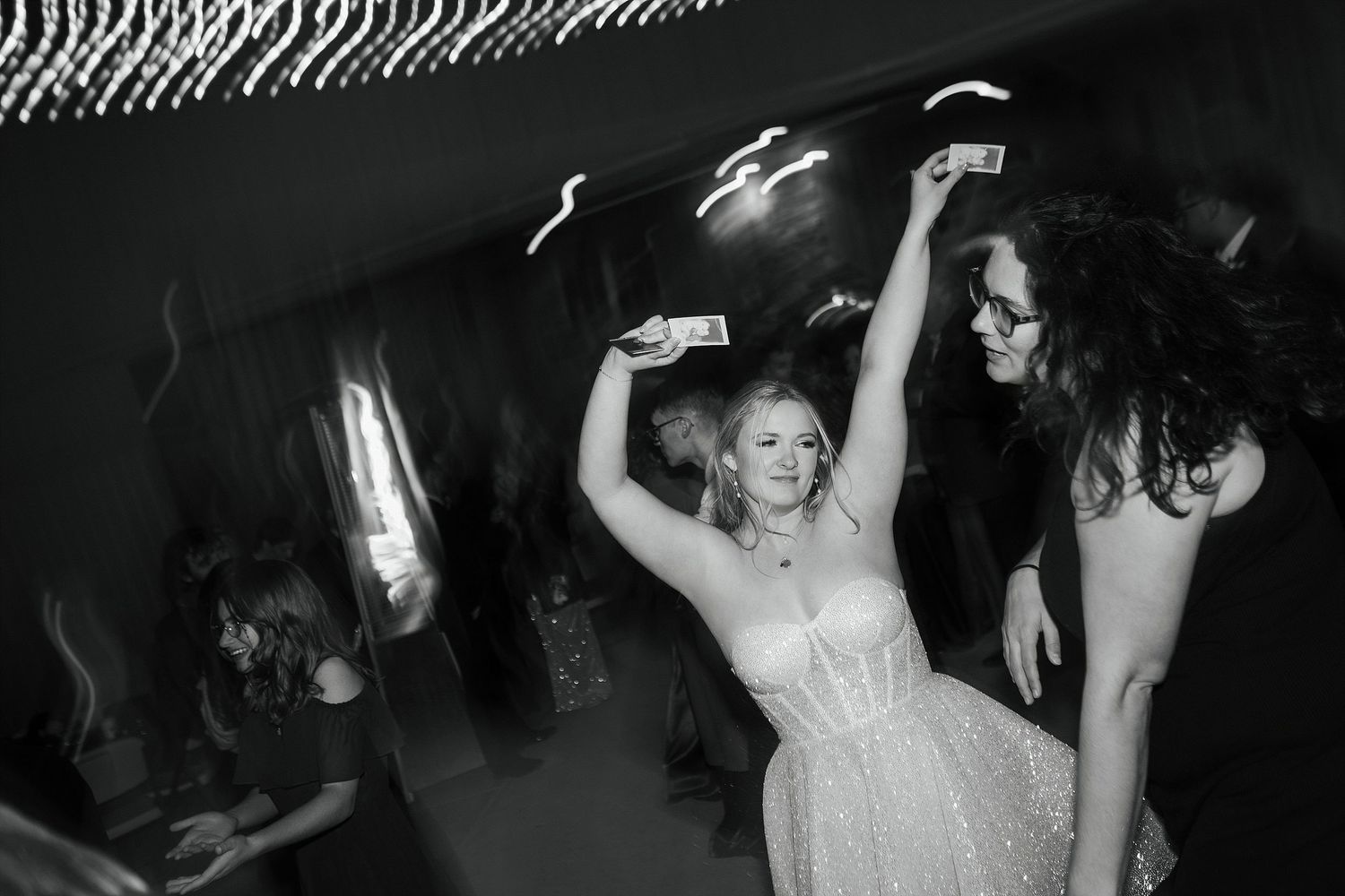 A black and white party scene shows people dancing with their arms raised in celebration at a festive gathering.