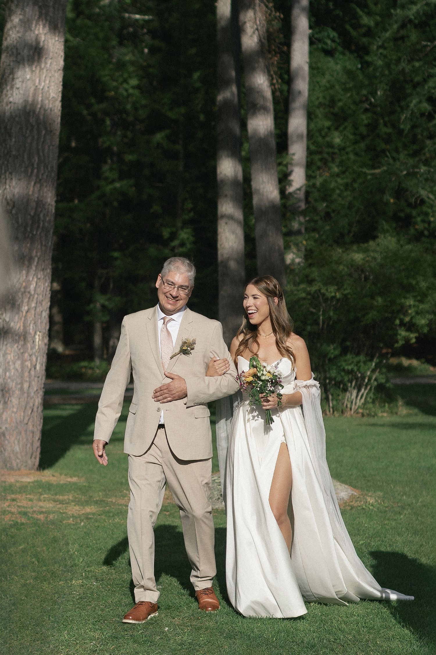 A couple walks down a garden path during their outdoor wedding ceremony surrounded by tall trees.