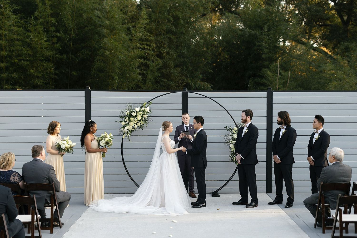 An intimate outdoor wedding ceremony takes place on a modern patio with a minimalist arch and grey backdrop wall.