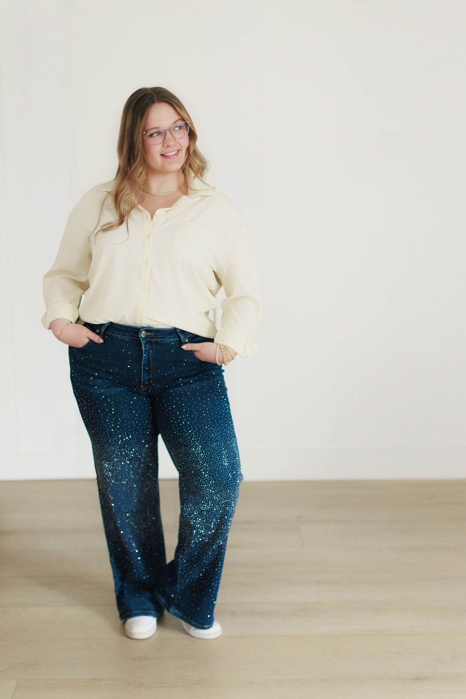 Woman in cream blouse and sparkly dark blue wide-leg jeans with white sneakers and glasses, posing confidently.