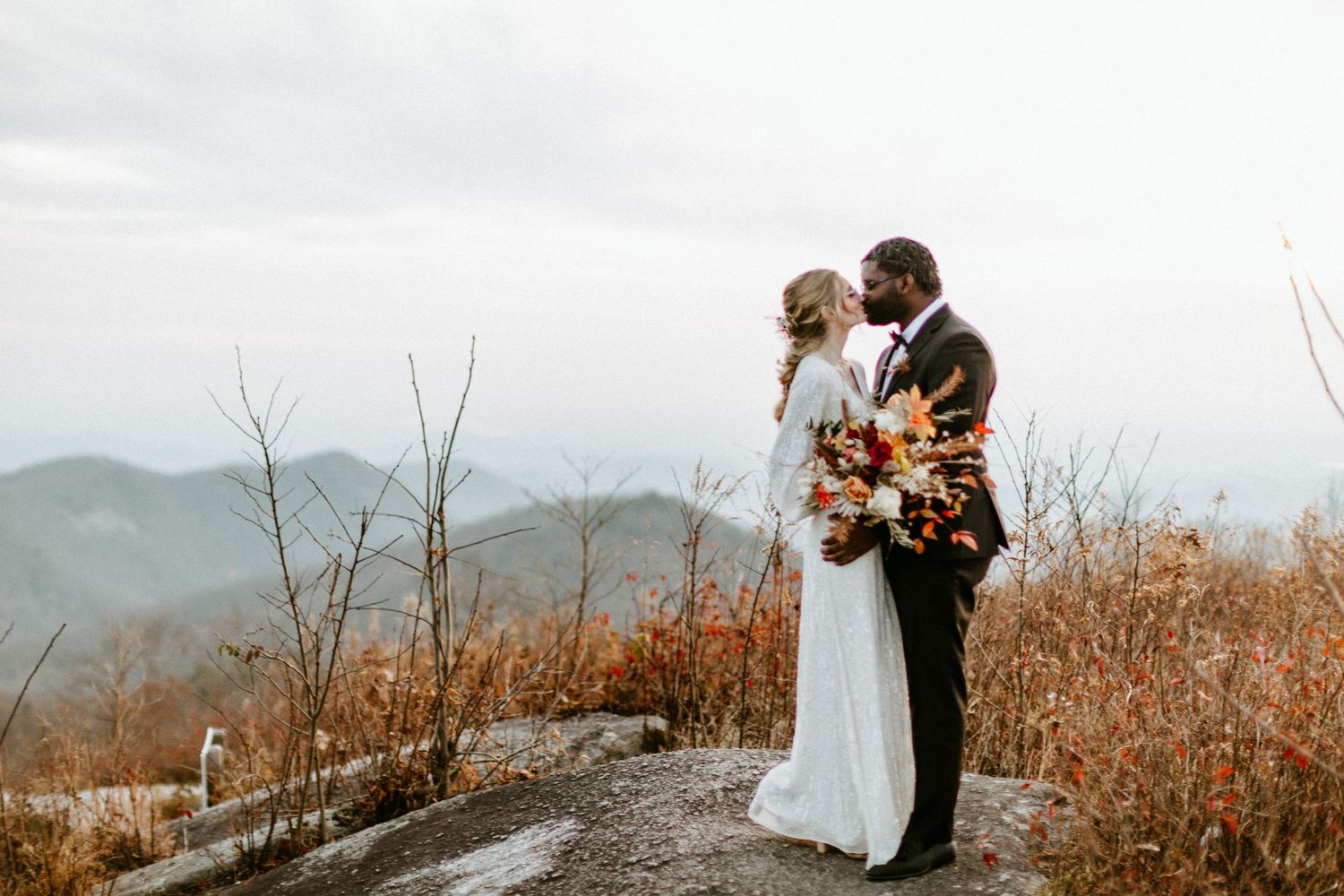 A couple stands on a mountain overlook embracing with fall foliage and misty peaks in the background.