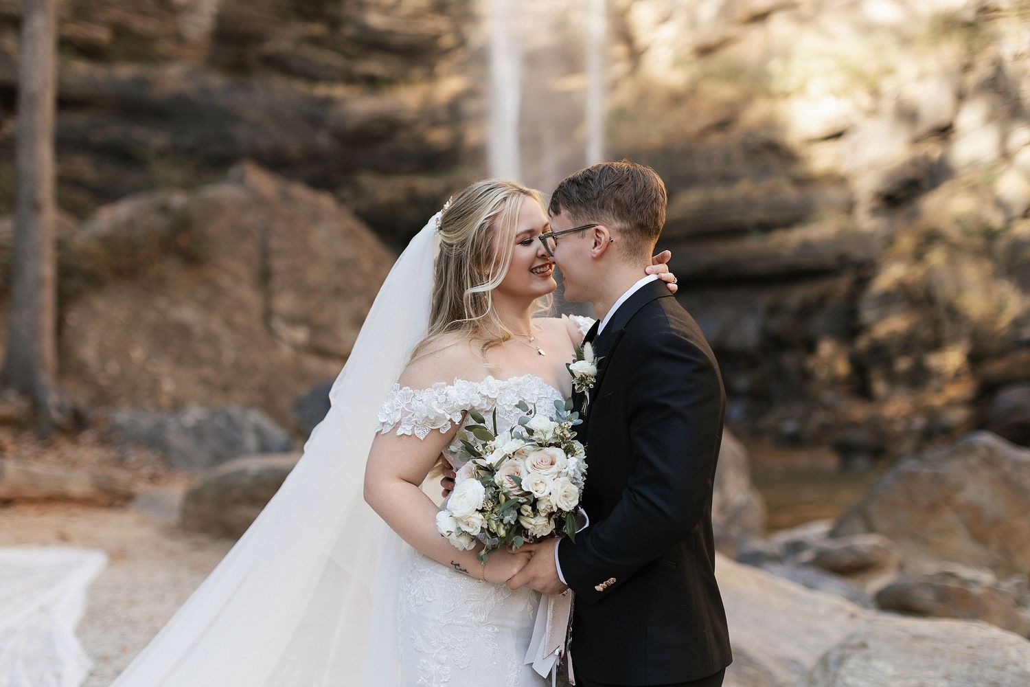 A romantic wedding couple embraces in front of a scenic waterfall backdrop while holding an elegant white floral bouquet.
