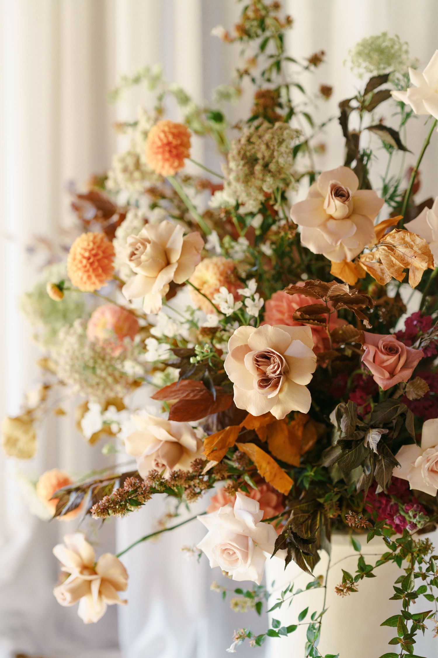 Lush autumn floral arrangement featuring peach roses, dahlias, and dried botanicals in warm earth tones against a white backdrop.
