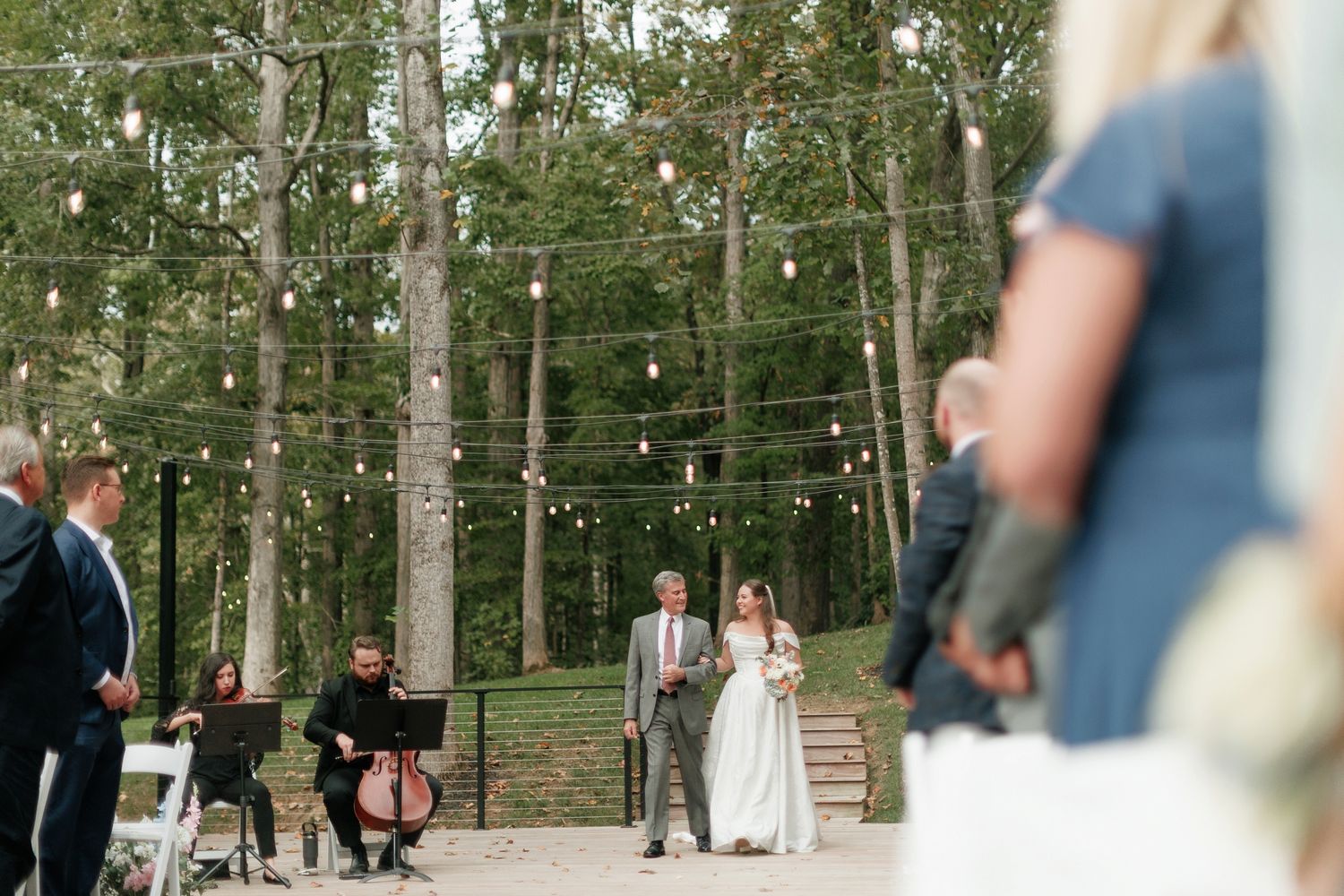 Outdoor wedding ceremony in a forest with string lights, couple at the altar, guests seated on white chairs.