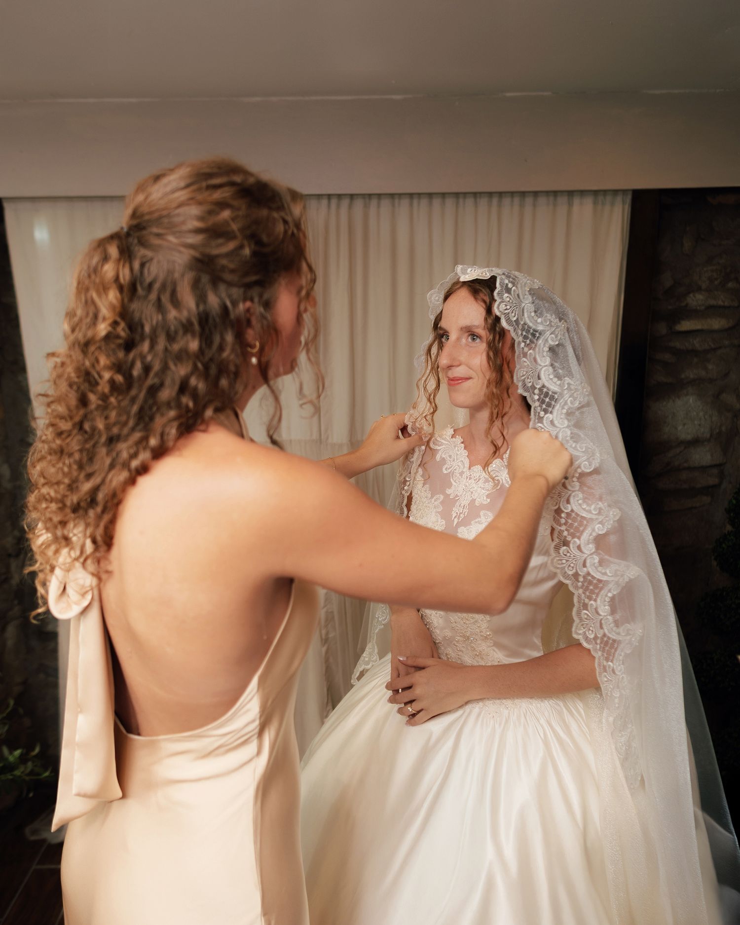 A bridesmaid adjusts the lace veil of a bride in a white wedding gown before the ceremony.