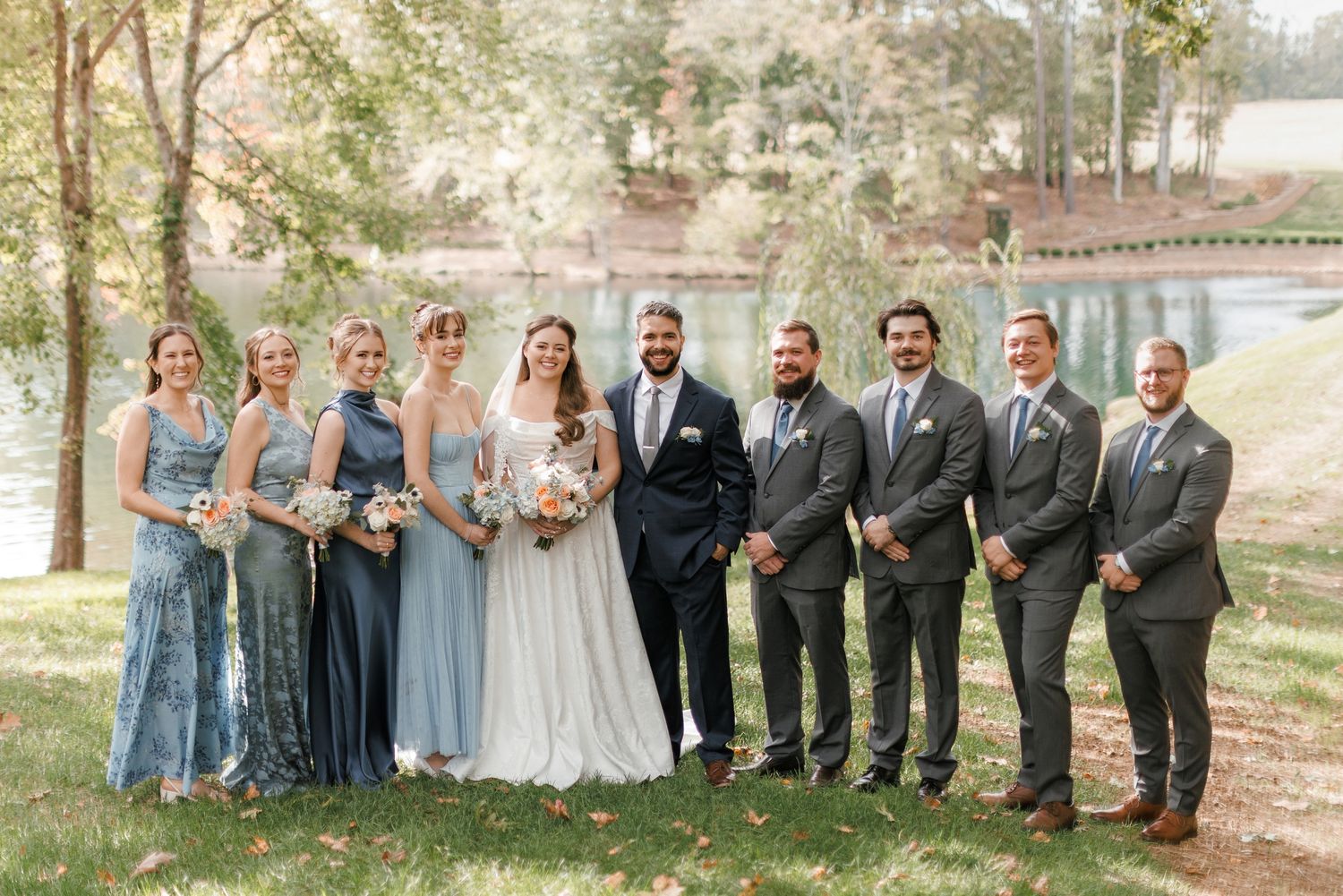 Wedding party poses outdoors by a lake, bridesmaids in blue gowns and groomsmen in gray suits.