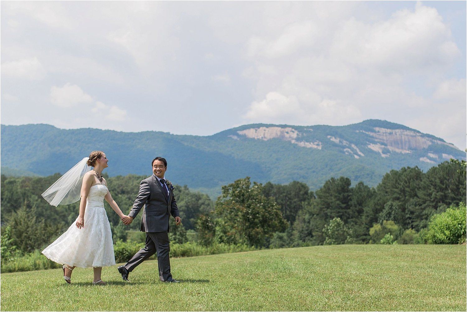 Newlyweds walking hand in hand on grassy hill with mountain backdrop.