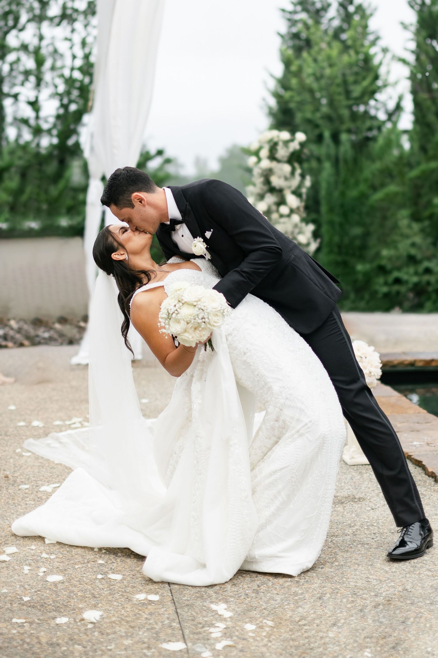 Classic wedding dip pose on stone patio with white floral arrangements and greenery in background.