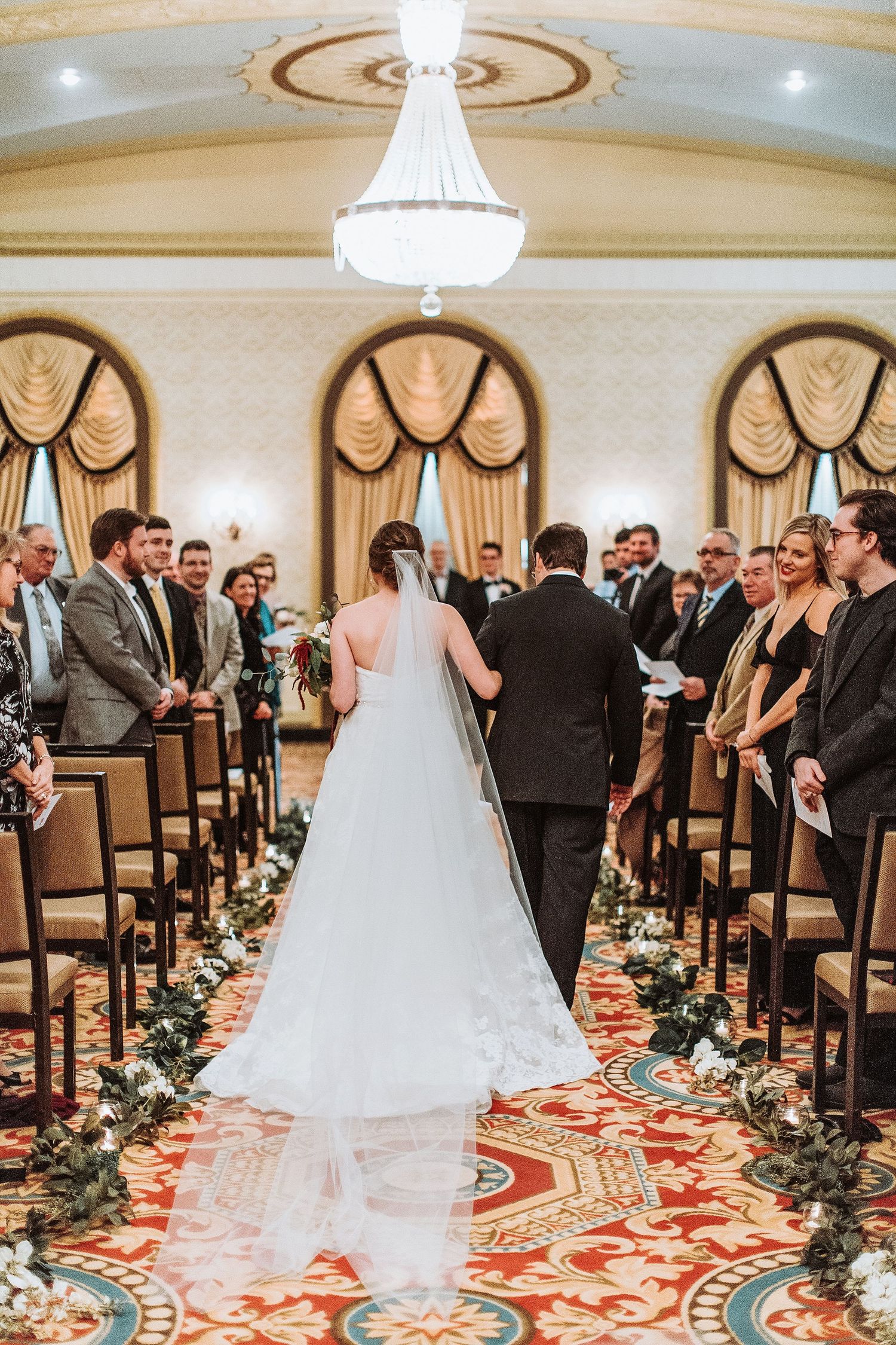 A wedding ceremony in an elegant ballroom with chandelier and floral-lined aisle.