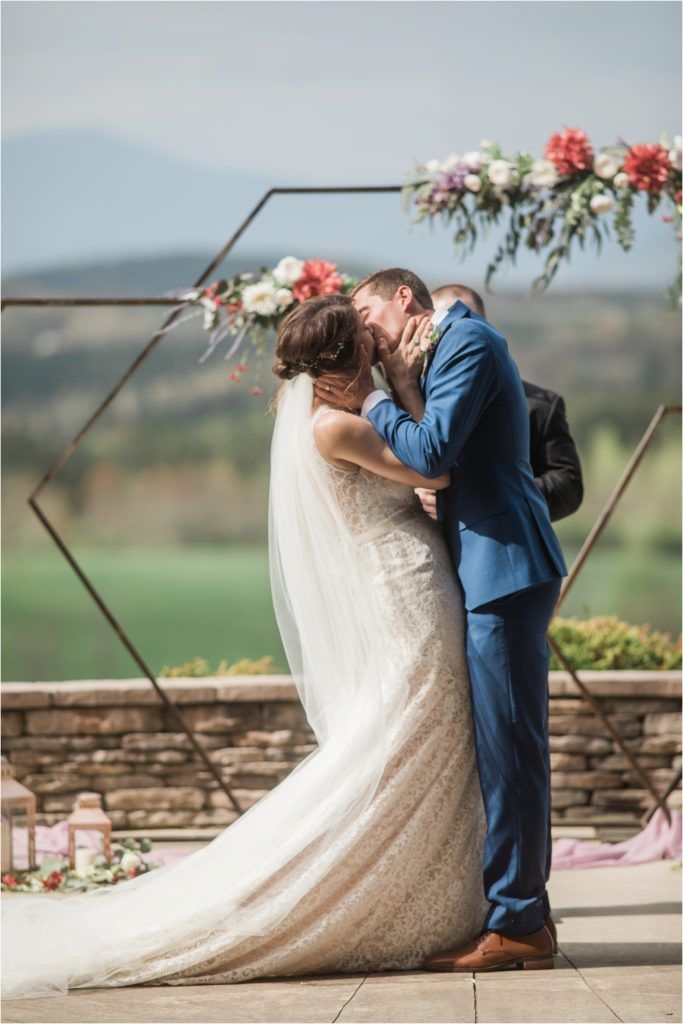 Couple sharing their first kiss under a floral hexagon arch at an outdoor mountain venue.
