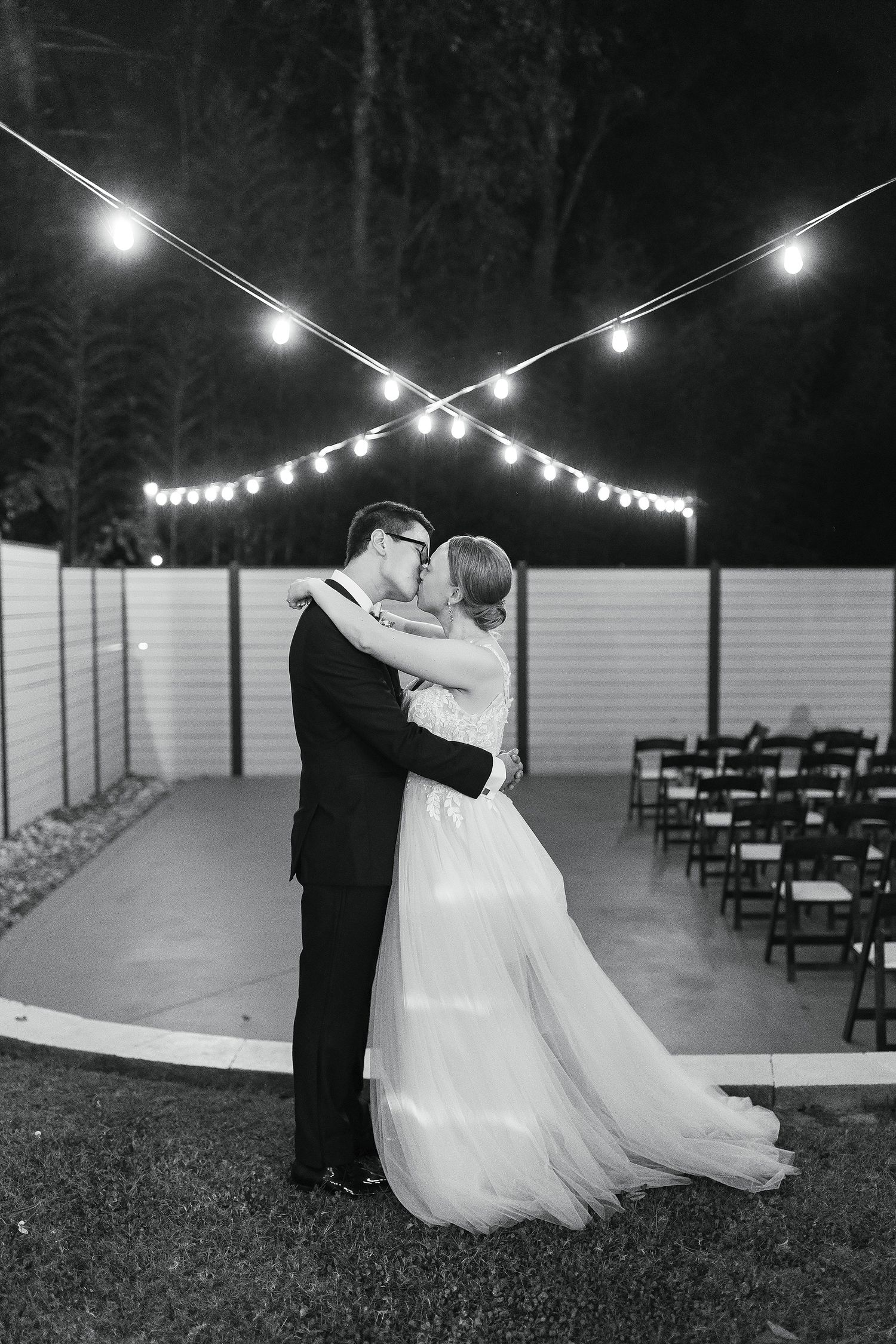 A romantic black and white outdoor wedding photo with string lights illuminating the evening ceremony space.
