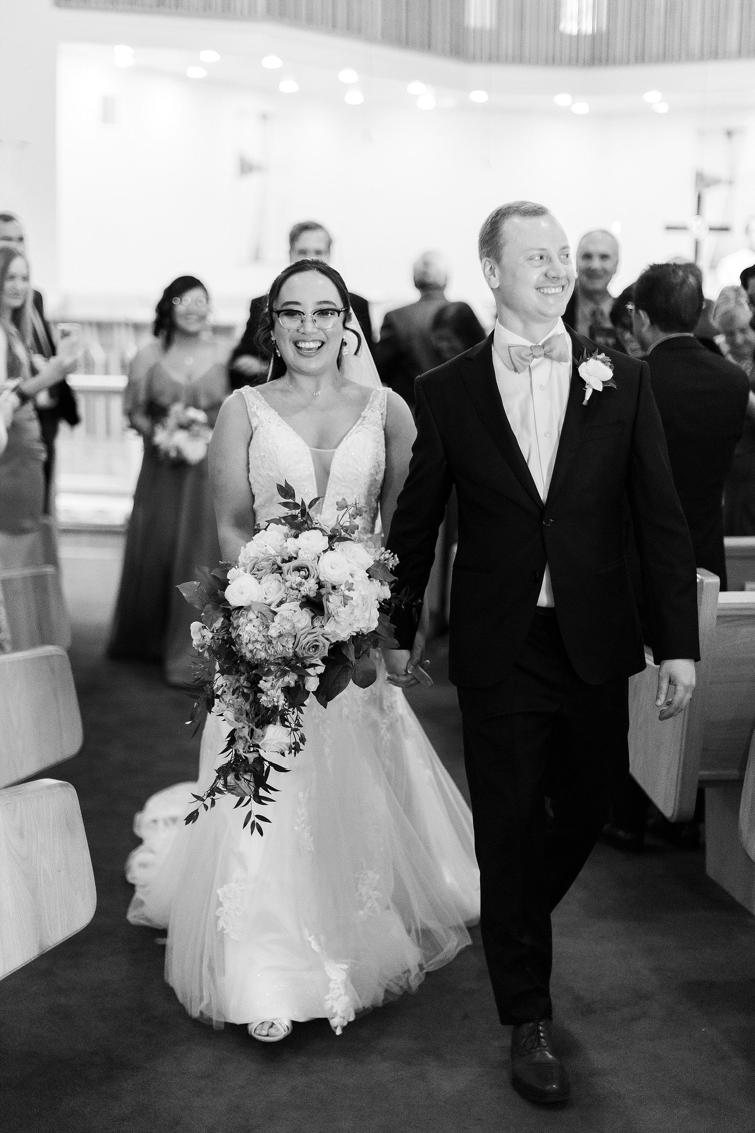 Bride in white lace gown and groom exit church ceremony with joyful expressions.