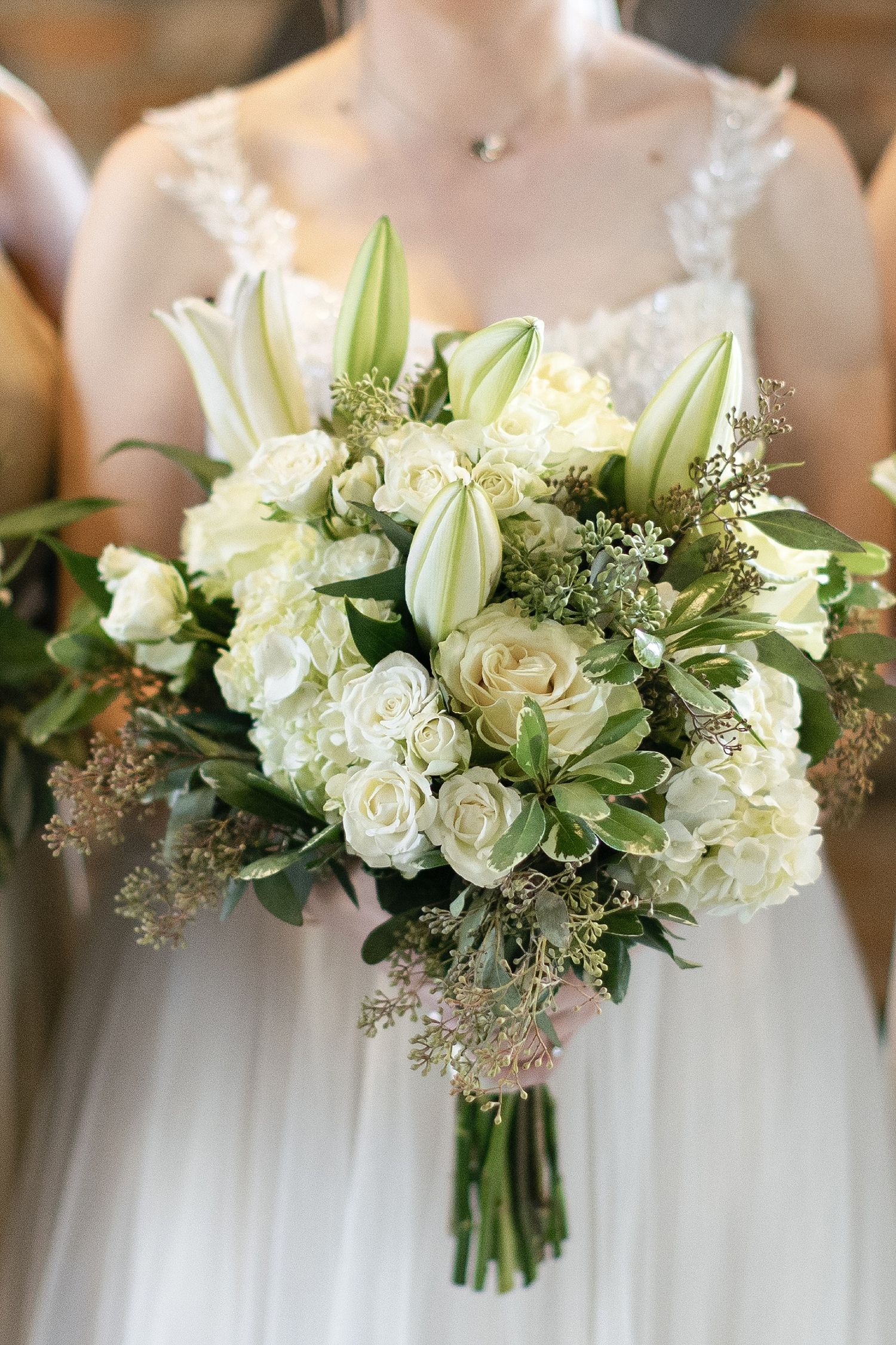 Elegant white wedding bouquet featuring lilies, roses, and greenery held in front of a lace dress.