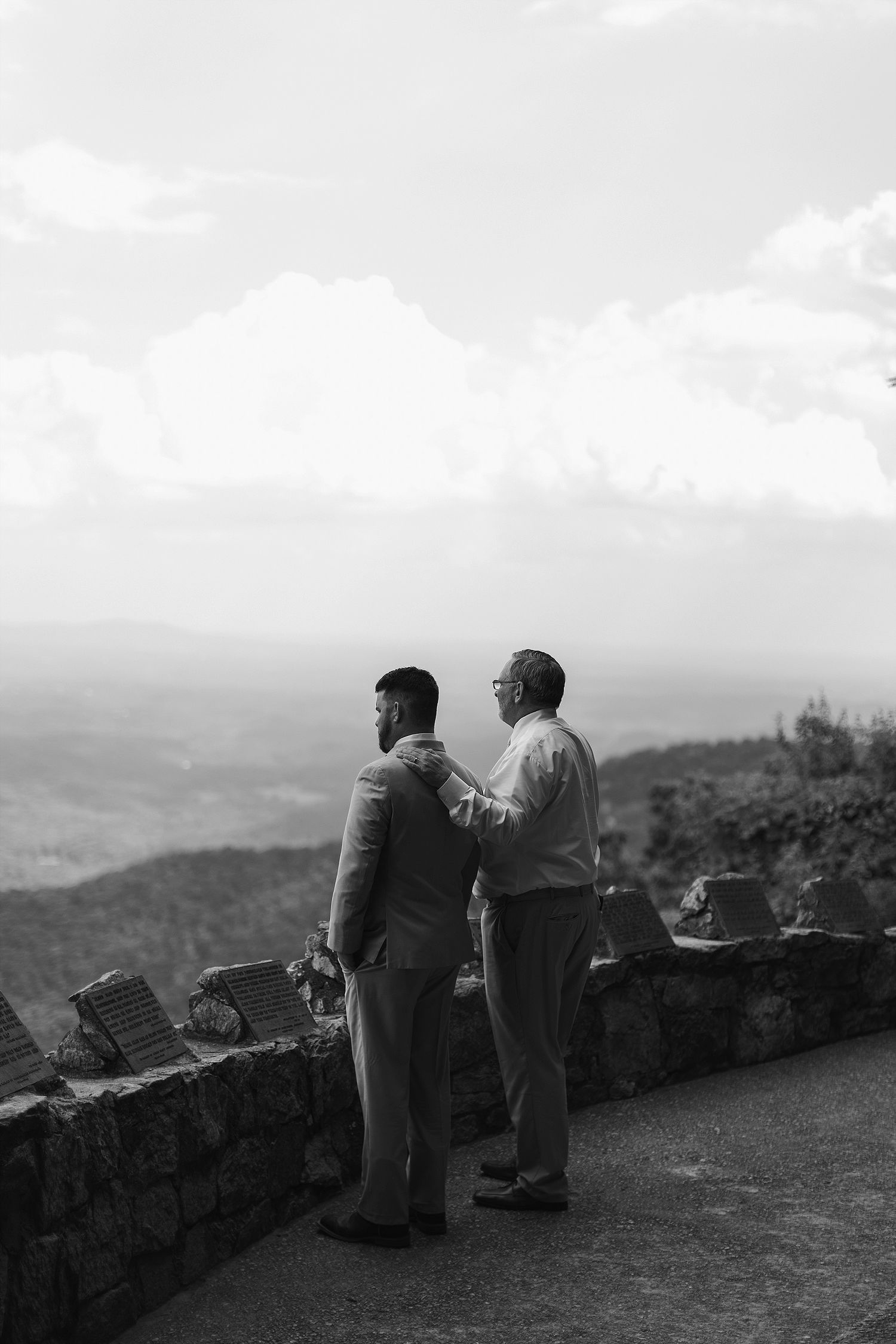 Two figures stand at a scenic mountain overlook with stone wall in black and white.