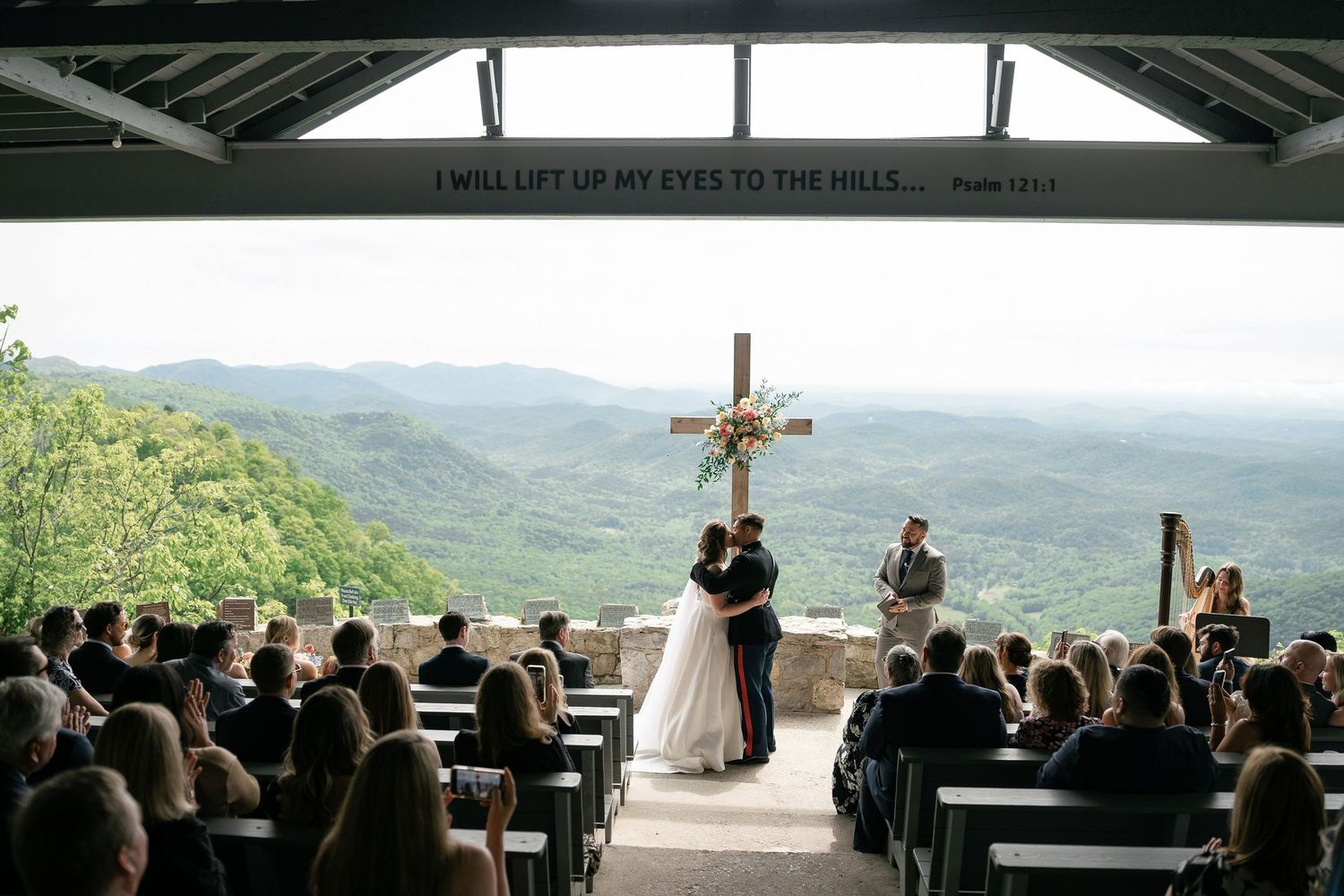 A mountain wedding ceremony takes place in an open-air chapel with panoramic views of rolling green hills beneath cloudy skies.