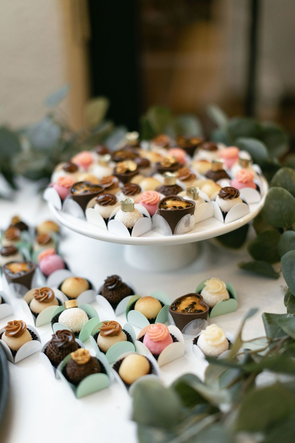 Tiered display of colorful chocolate truffles and petit fours decorated with eucalyptus leaves.