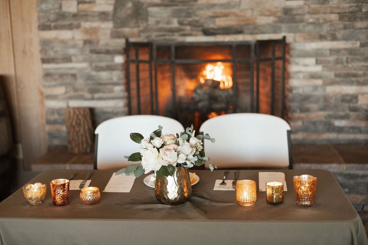 Romantic table setting with candlelight and floral centerpiece in front of a cozy stone fireplace.
