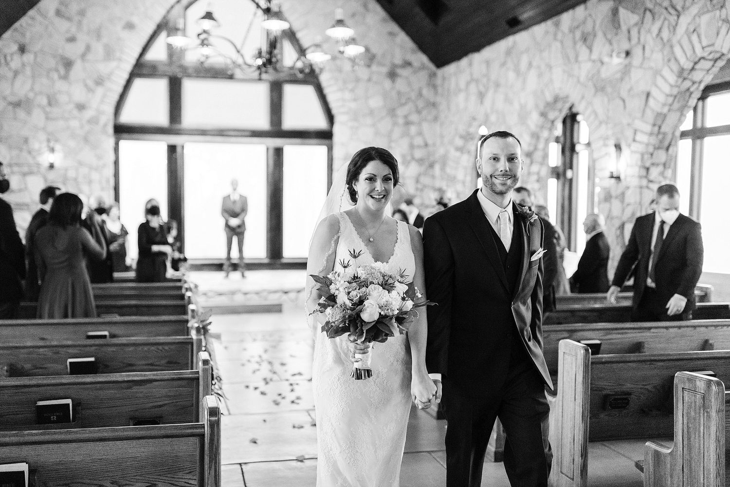 A couple walks down the aisle of a stone chapel with arched windows and wooden pews.