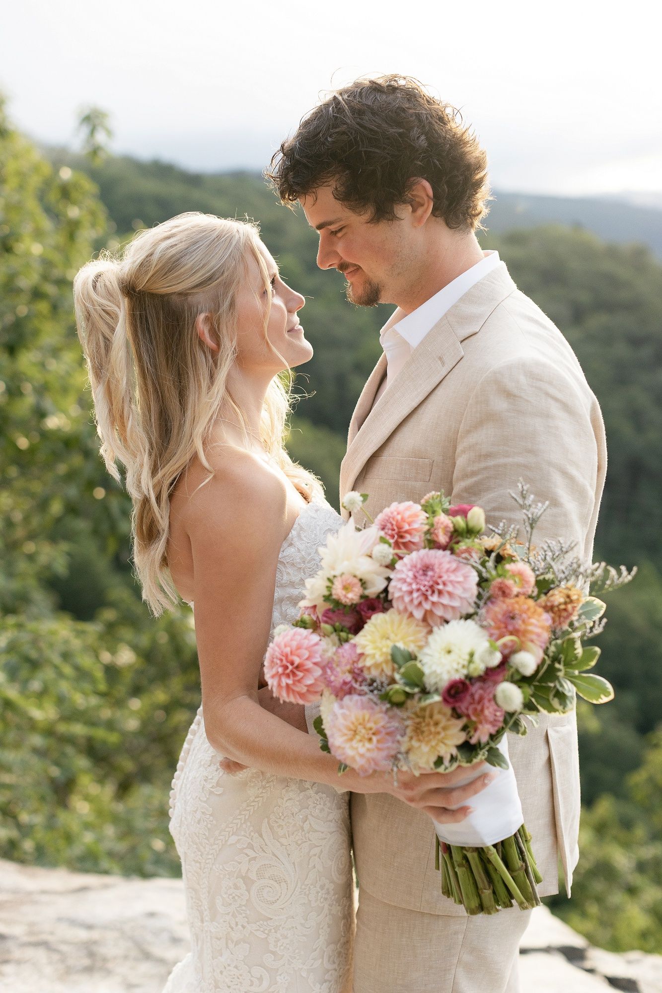 Wedding couple embracing outdoors with pink and white floral bouquet against scenic mountain backdrop.