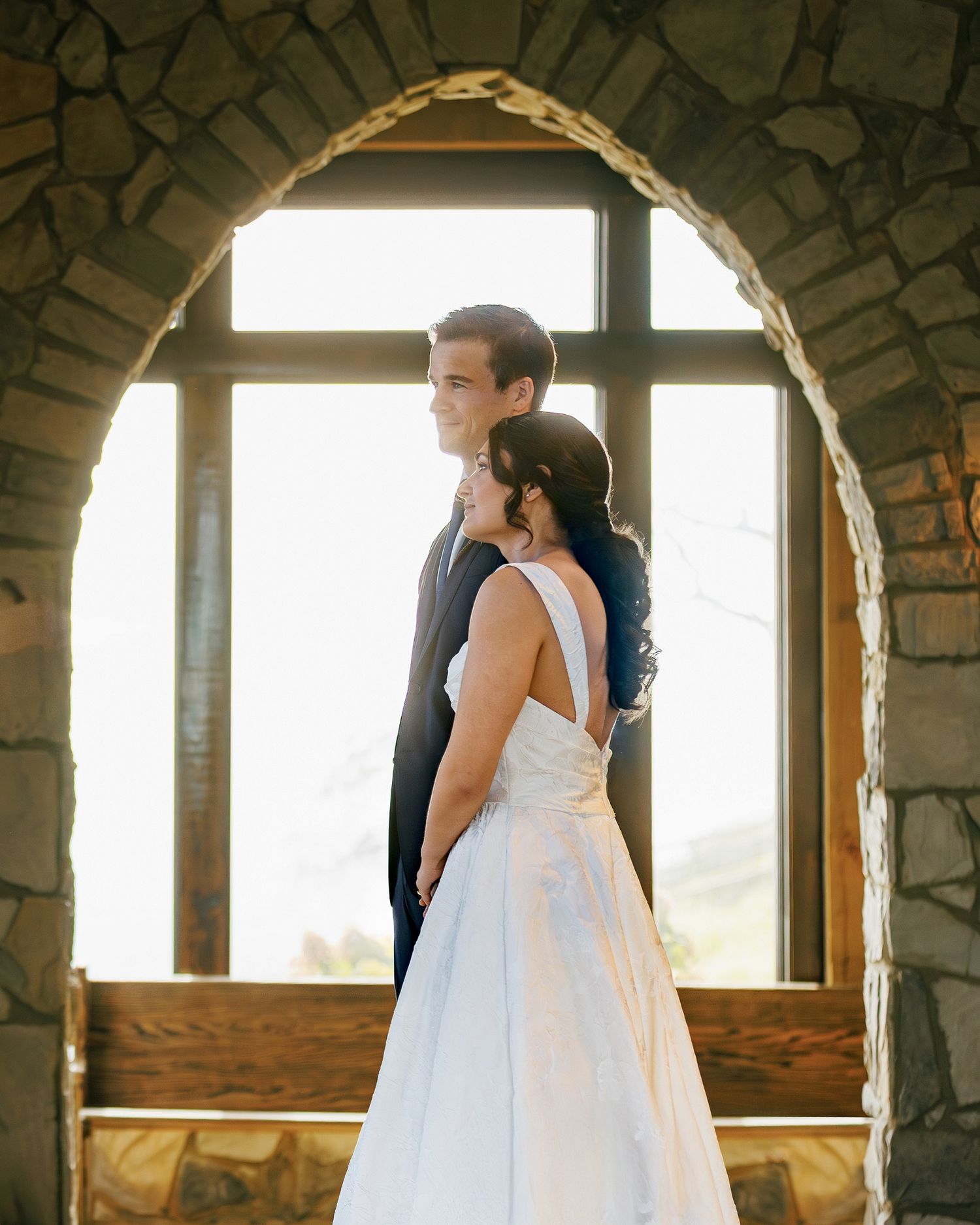 A couple in wedding attire stands silhouetted in a stone archway with sunlight streaming through windows behind them.