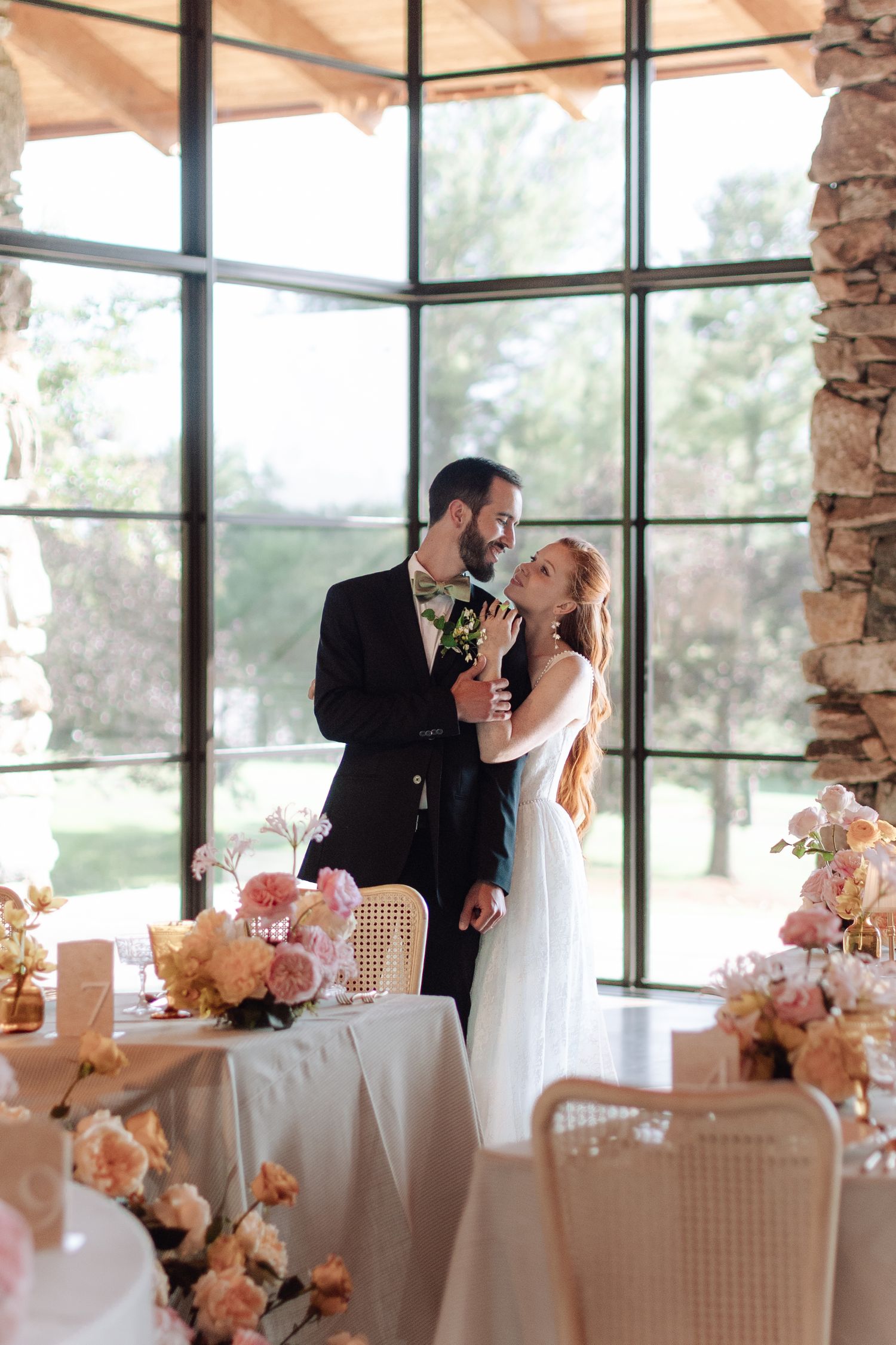 Bride and groom share a romantic moment at floral-decorated wedding reception with large windows and natural light.