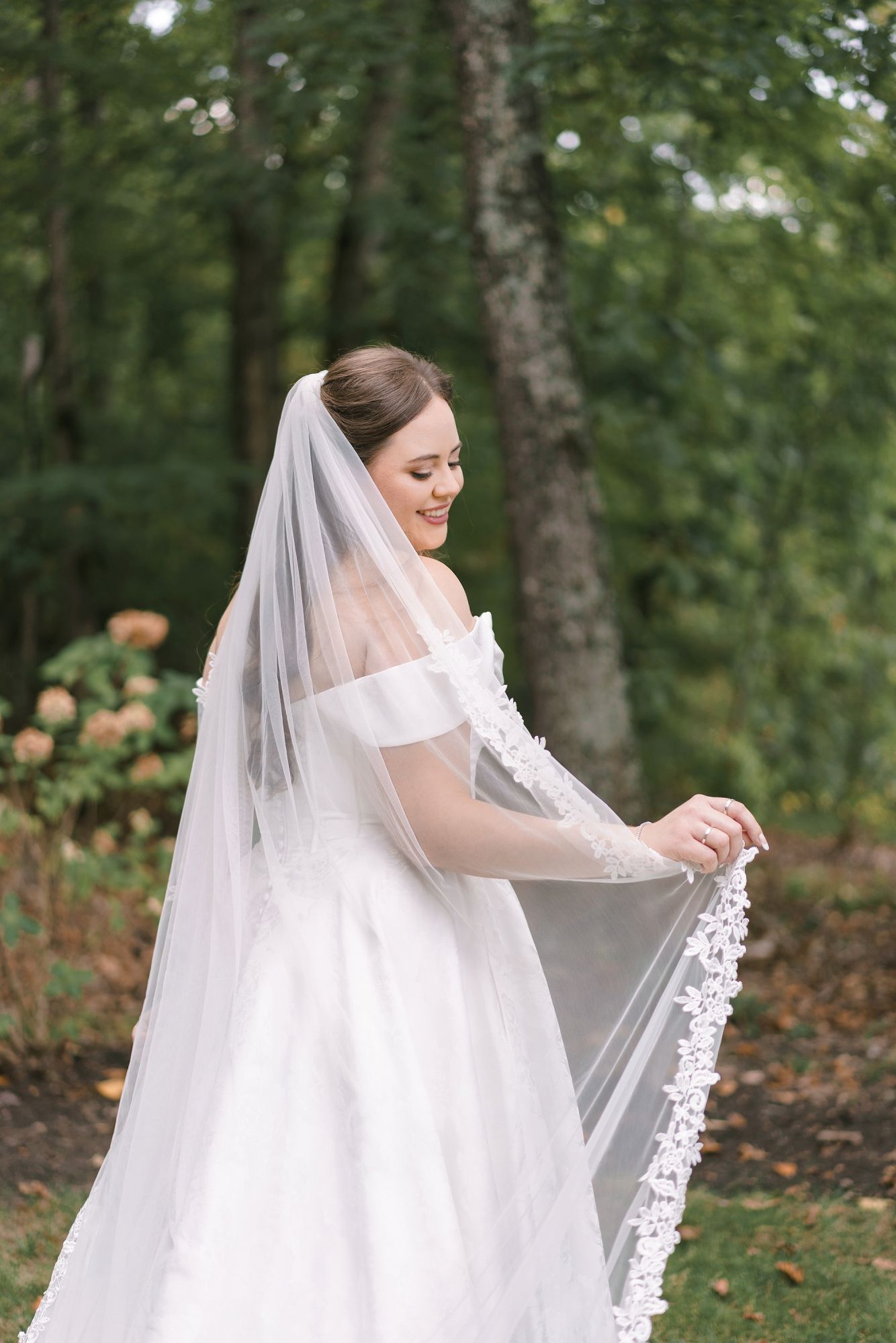 Smiling bride in white off-shoulder gown holding lace-trimmed veil in lush green garden setting.