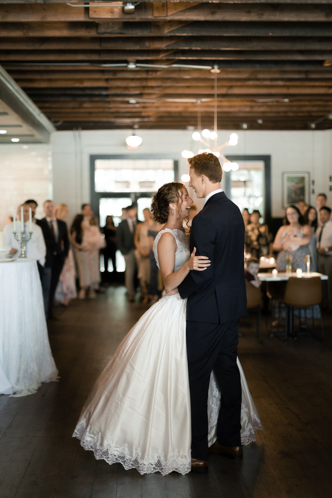 Bride and groom share first dance surrounded by guests at elegant indoor wedding reception.