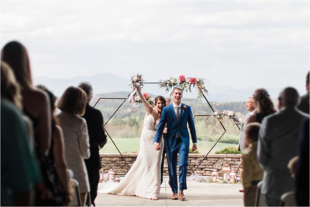 Newlyweds celebrating with raised arms at outdoor ceremony while guests applaud.