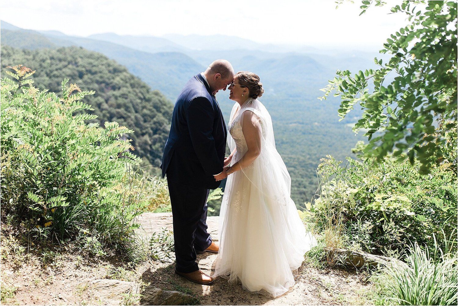 A couple shares an intimate moment on a mountain overlook surrounded by lush greenery.