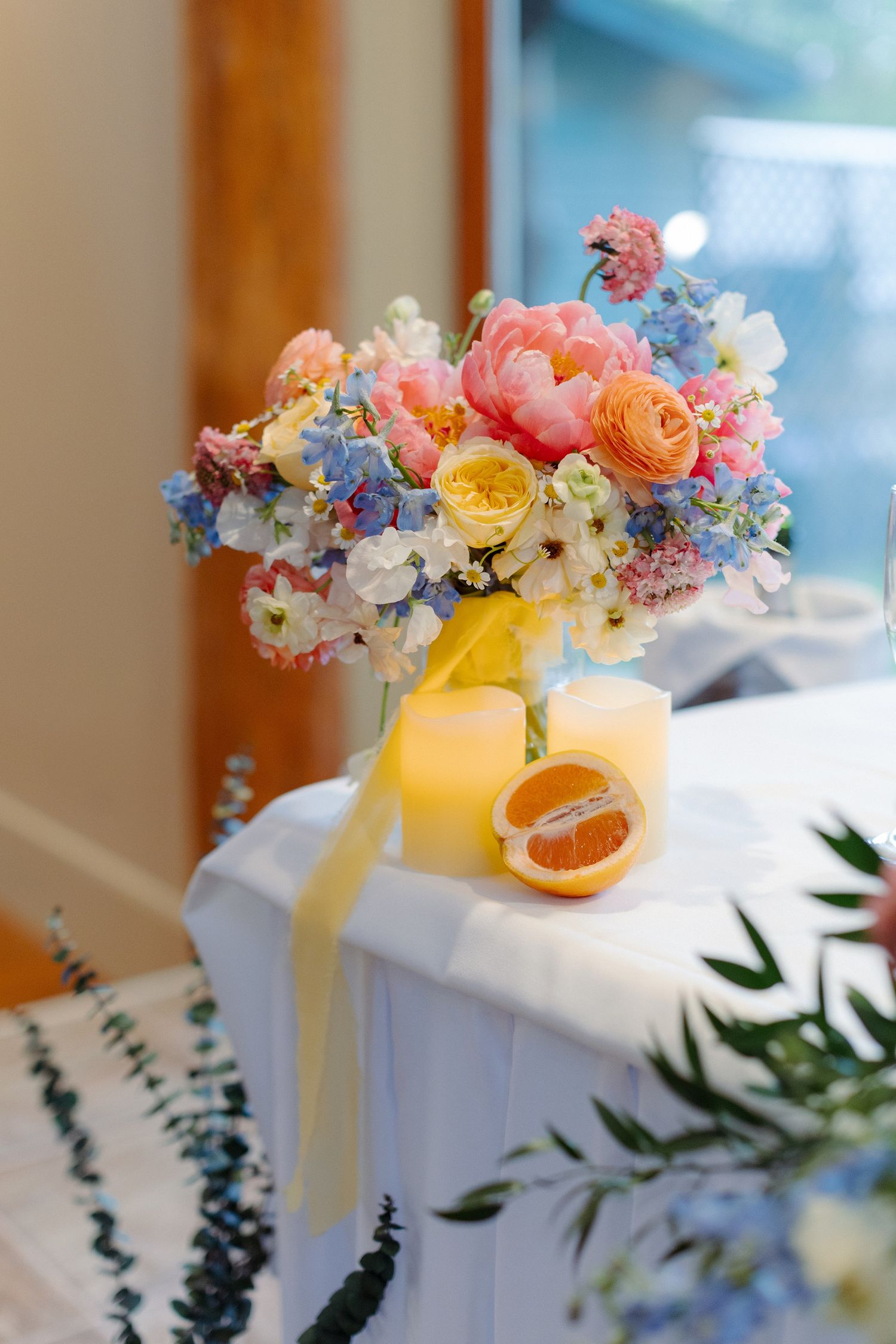 Beautiful floral arrangement with coral peonies, blue delphiniums and yellow ranunculus next to citrus drinks with ribbons.