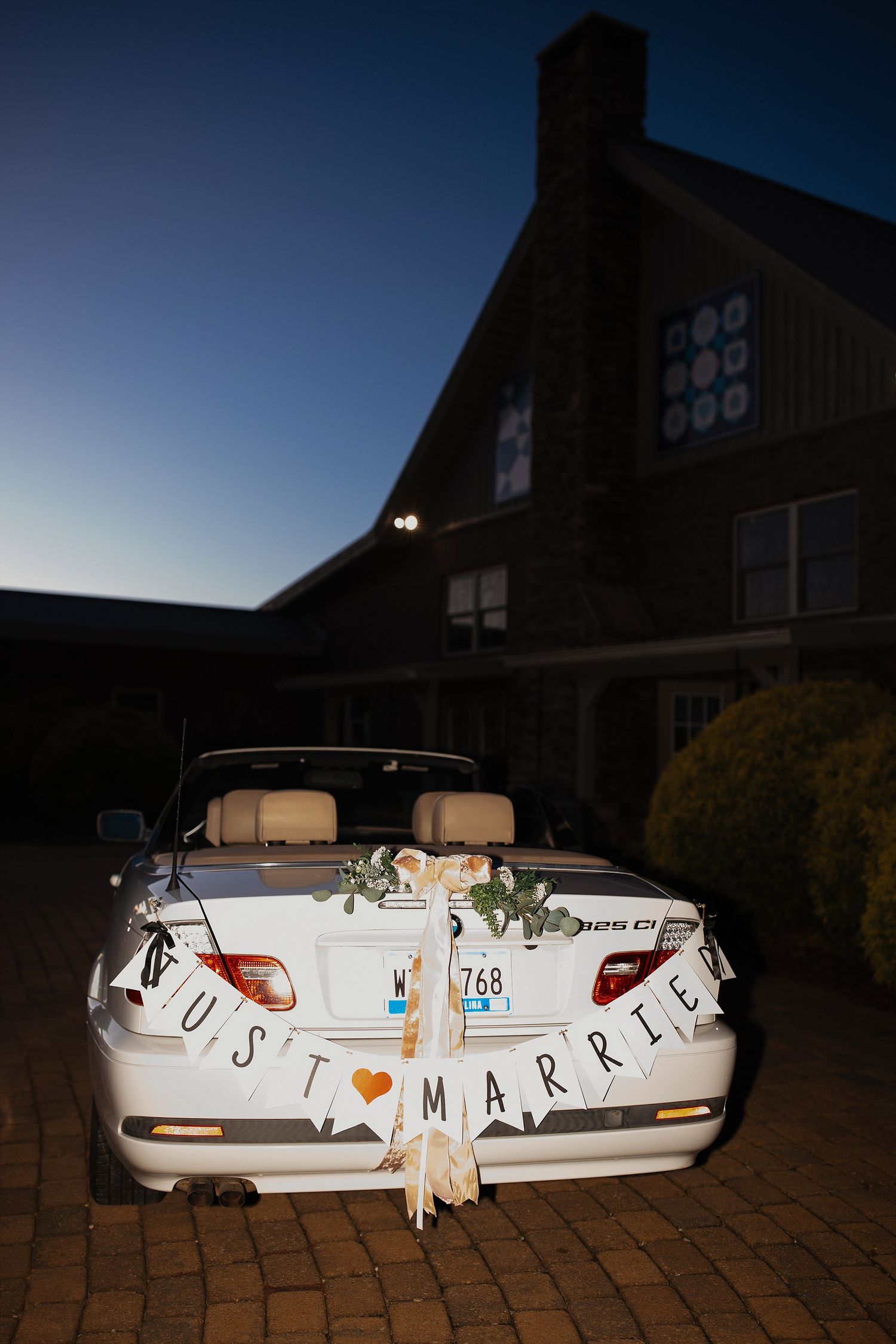 A white convertible wedding car decorated with 'Just Married' signage parked in front of a brick building at dusk.