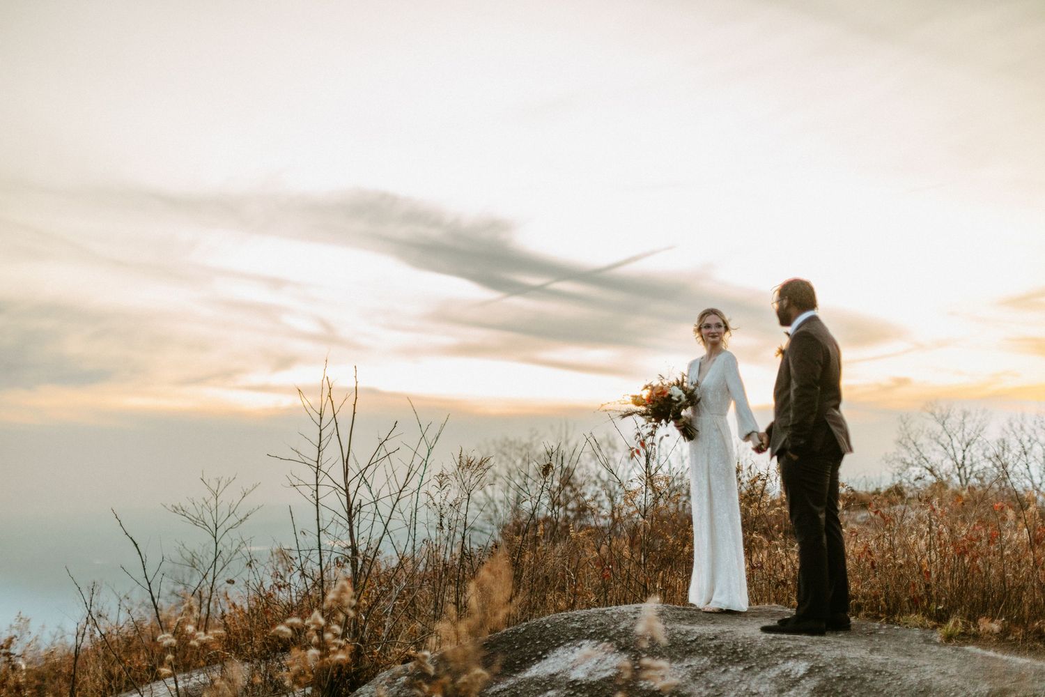 A couple enjoys a golden sunset moment while standing on a rocky mountain overlook.