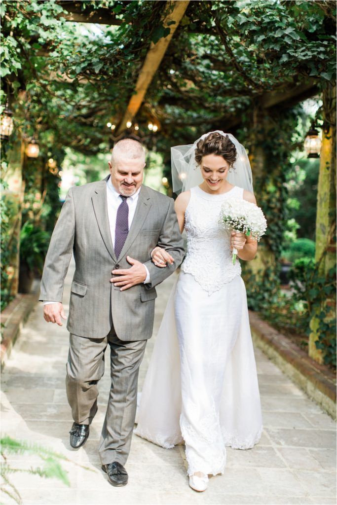 Bride in white lace gown walking down garden path.