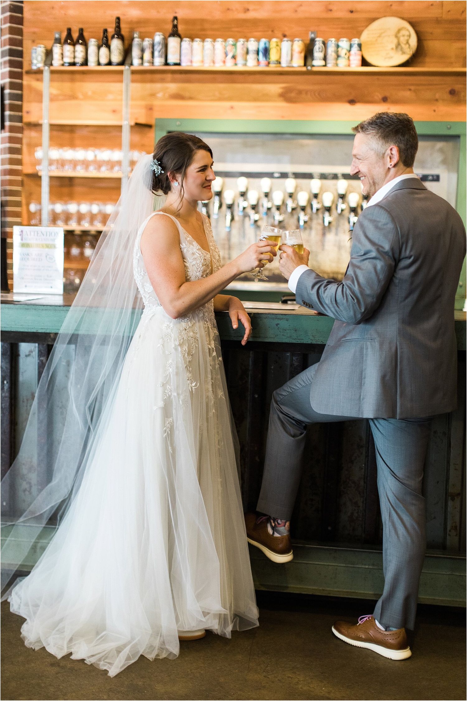Wedding couple shares drinks at a wooden bar with bottles displayed on shelves.