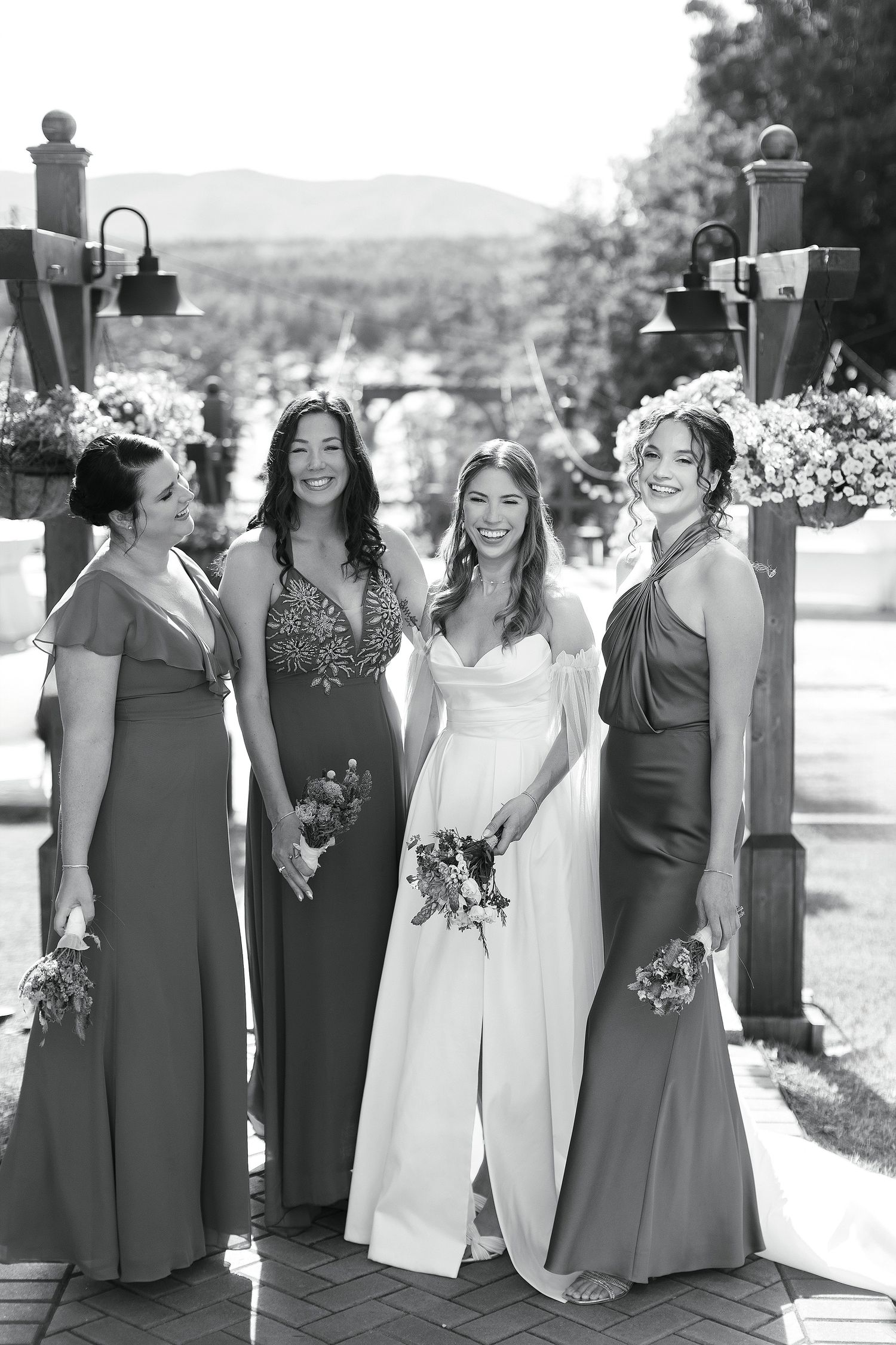 A bridal party in coordinating dresses poses together under wooden posts with floral arrangements.