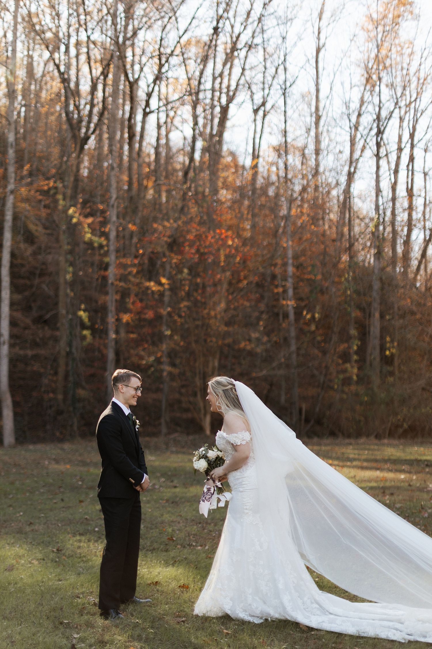 A couple in wedding attire stands in a clearing with fall trees and late afternoon sunlight in the background.