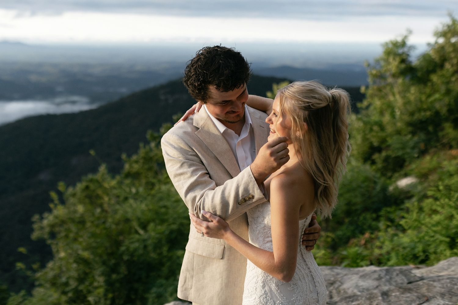 A couple in wedding attire share an intimate moment on a mountaintop overlook with scenic valley views behind them.