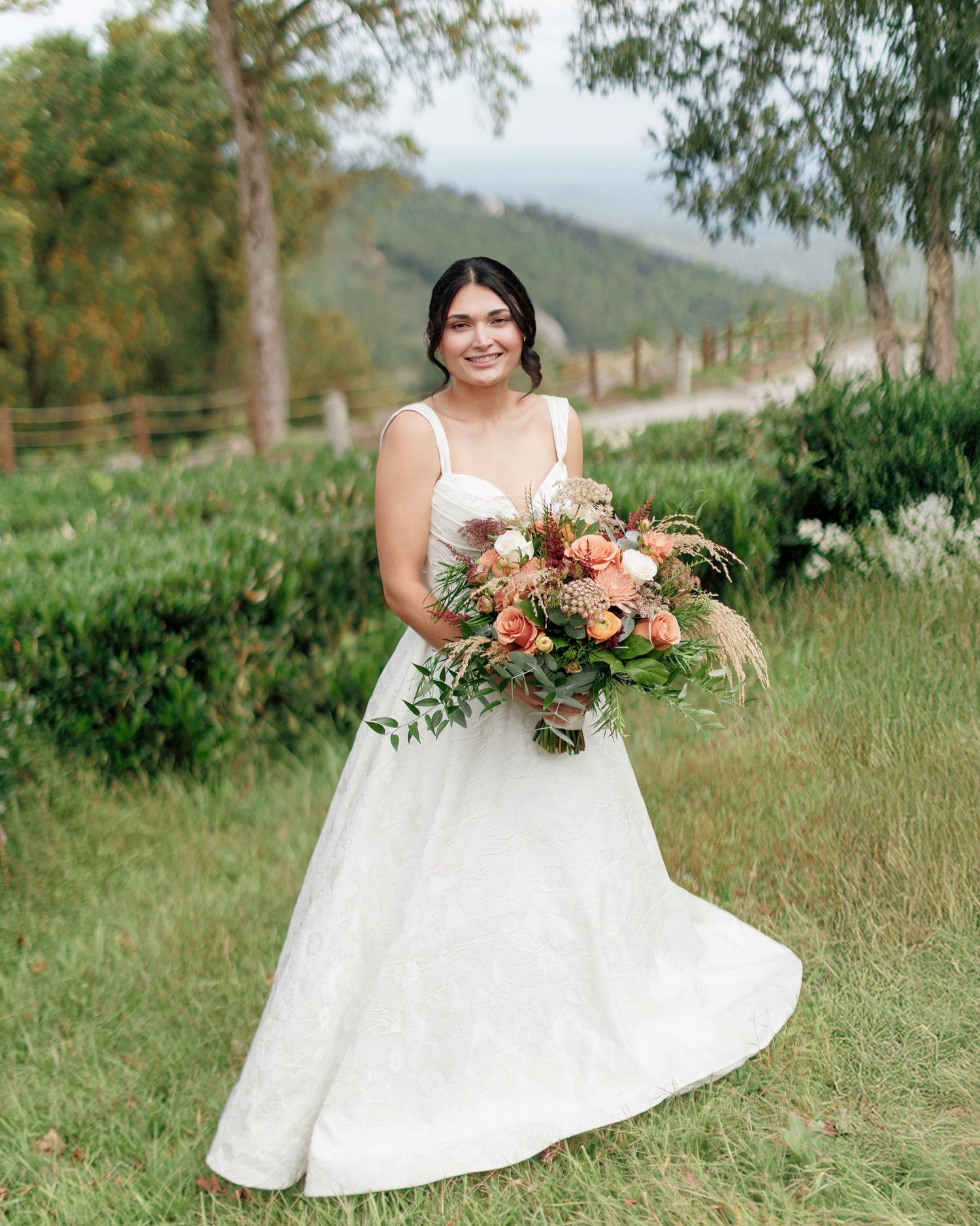 A bride in a flowing white dress holds a vibrant coral and peach floral bouquet in an outdoor garden setting.
