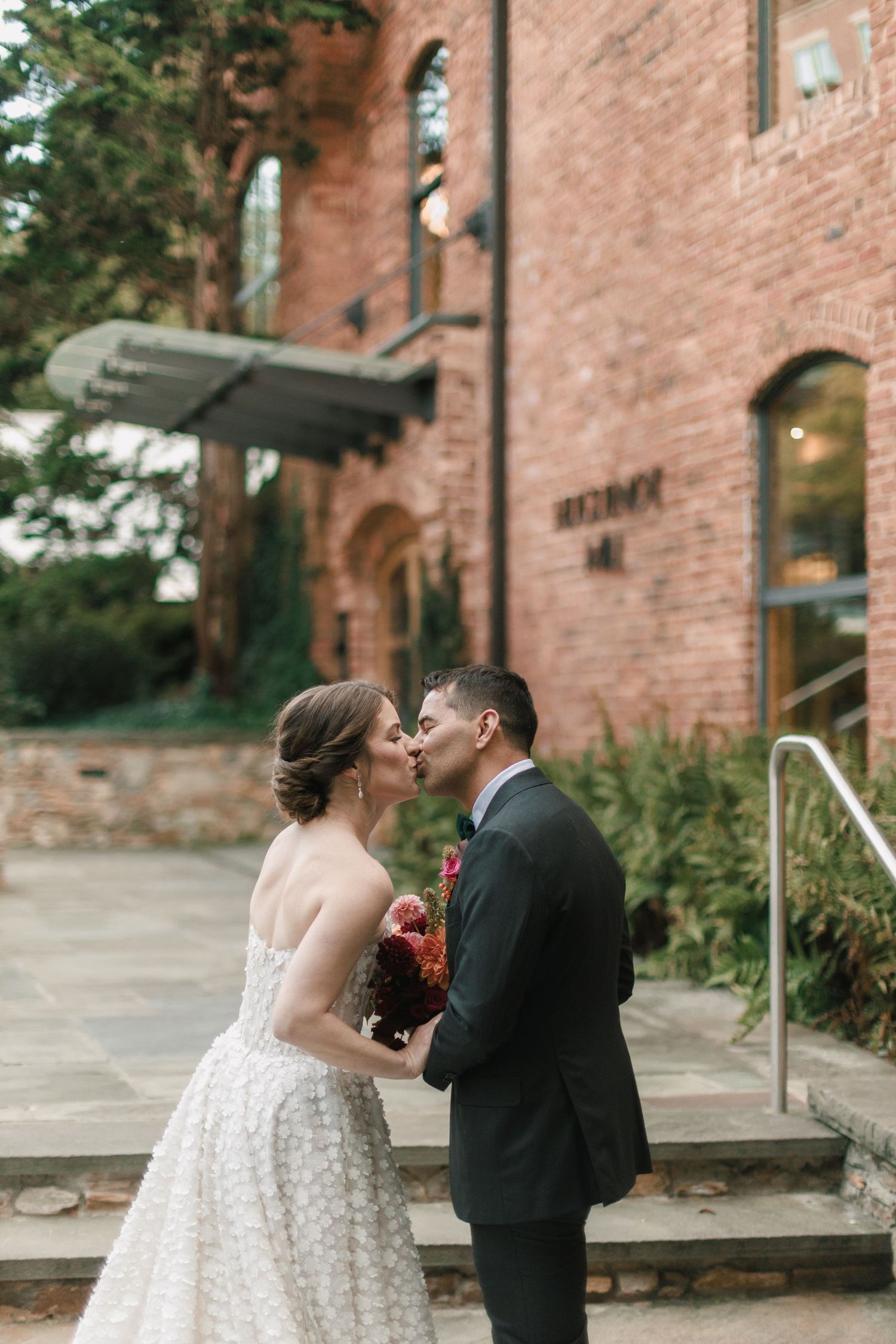 Bride and groom share a romantic kiss outside a rustic brick wedding venue, bride holding deep red bouquet.