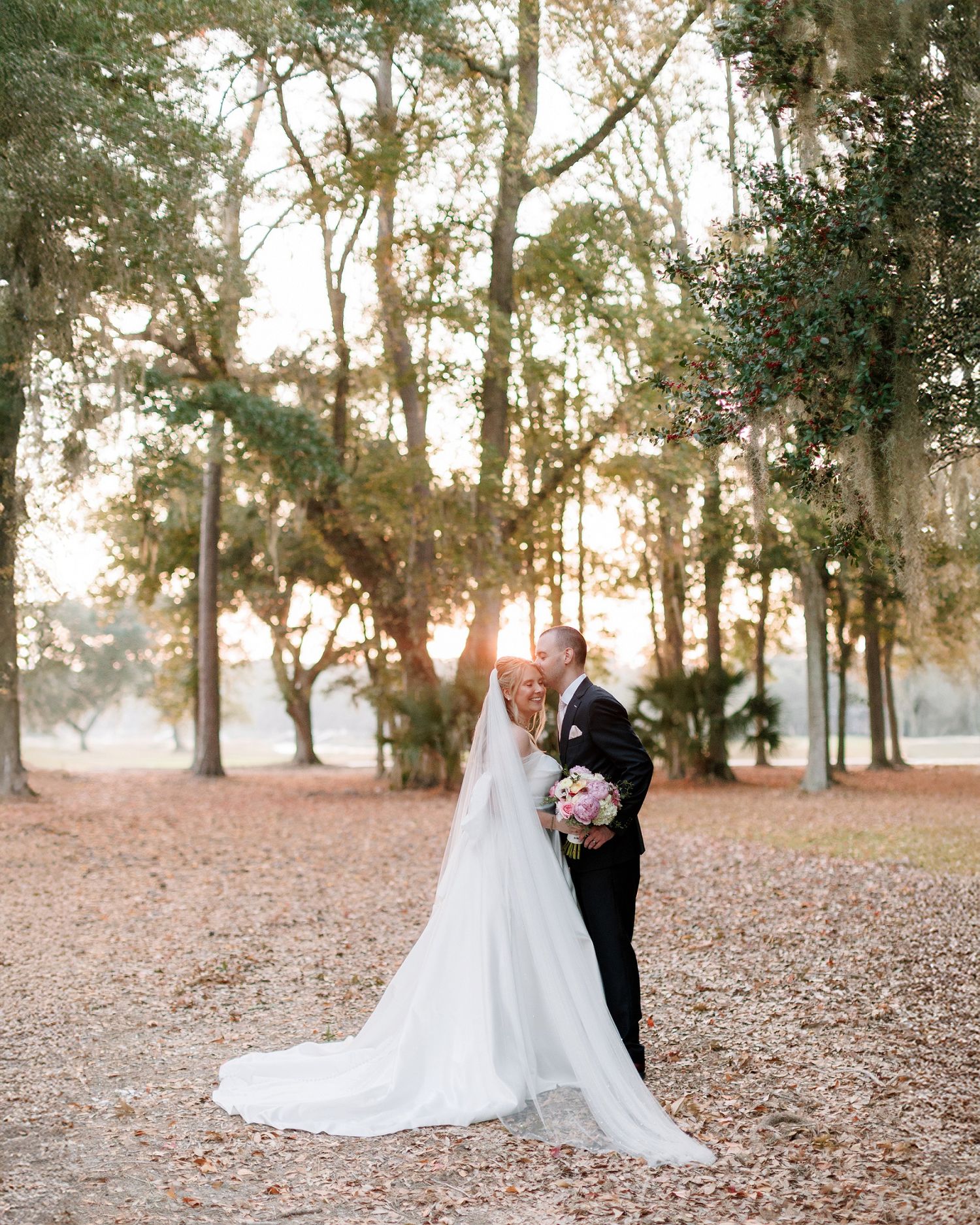 A romantic wedding couple dances together on fallen autumn leaves in a sunlit wooded area.