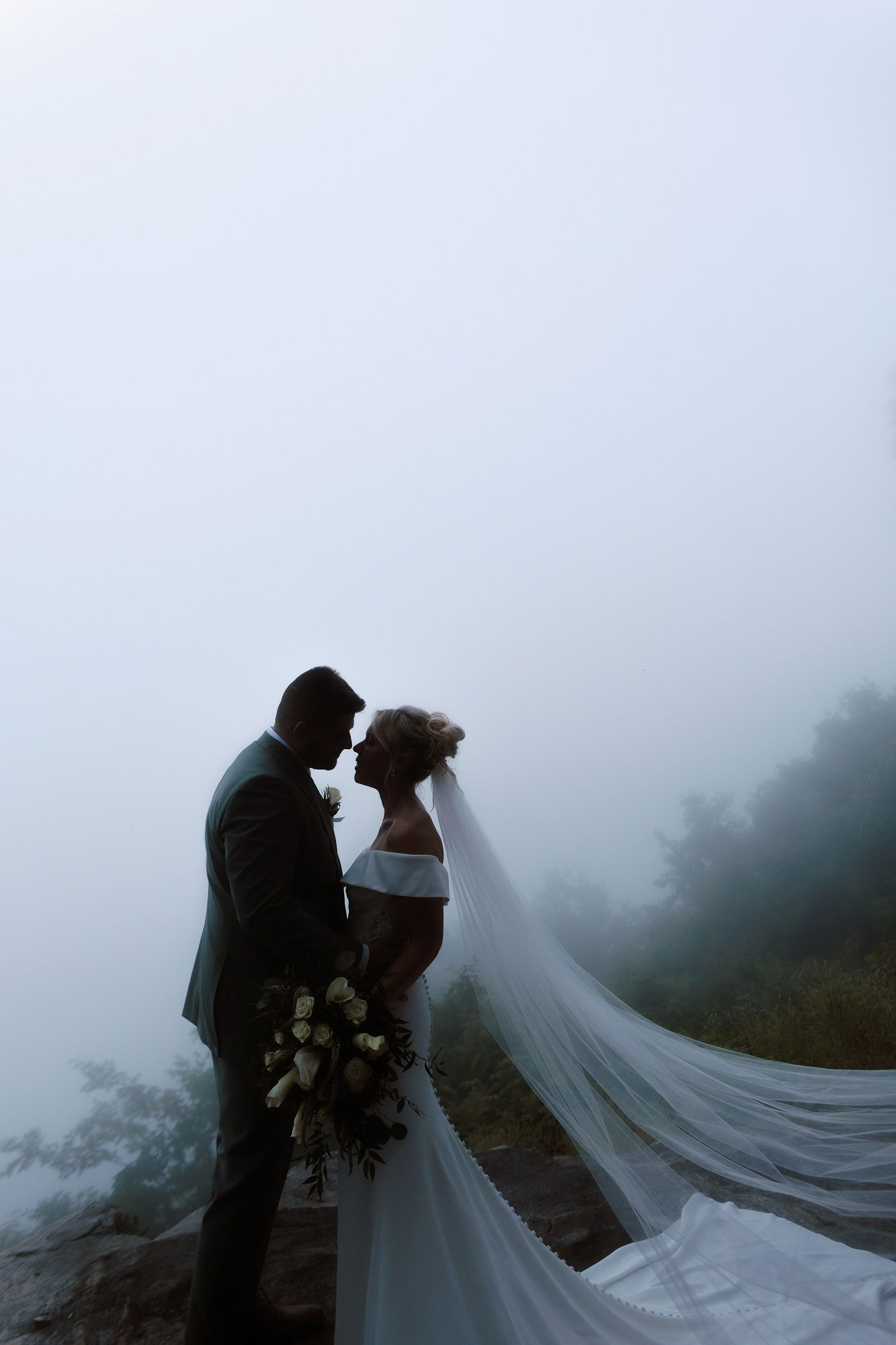 Silhouette of bride and groom sharing a tender moment in dense mountain fog with veil billowing in wind.